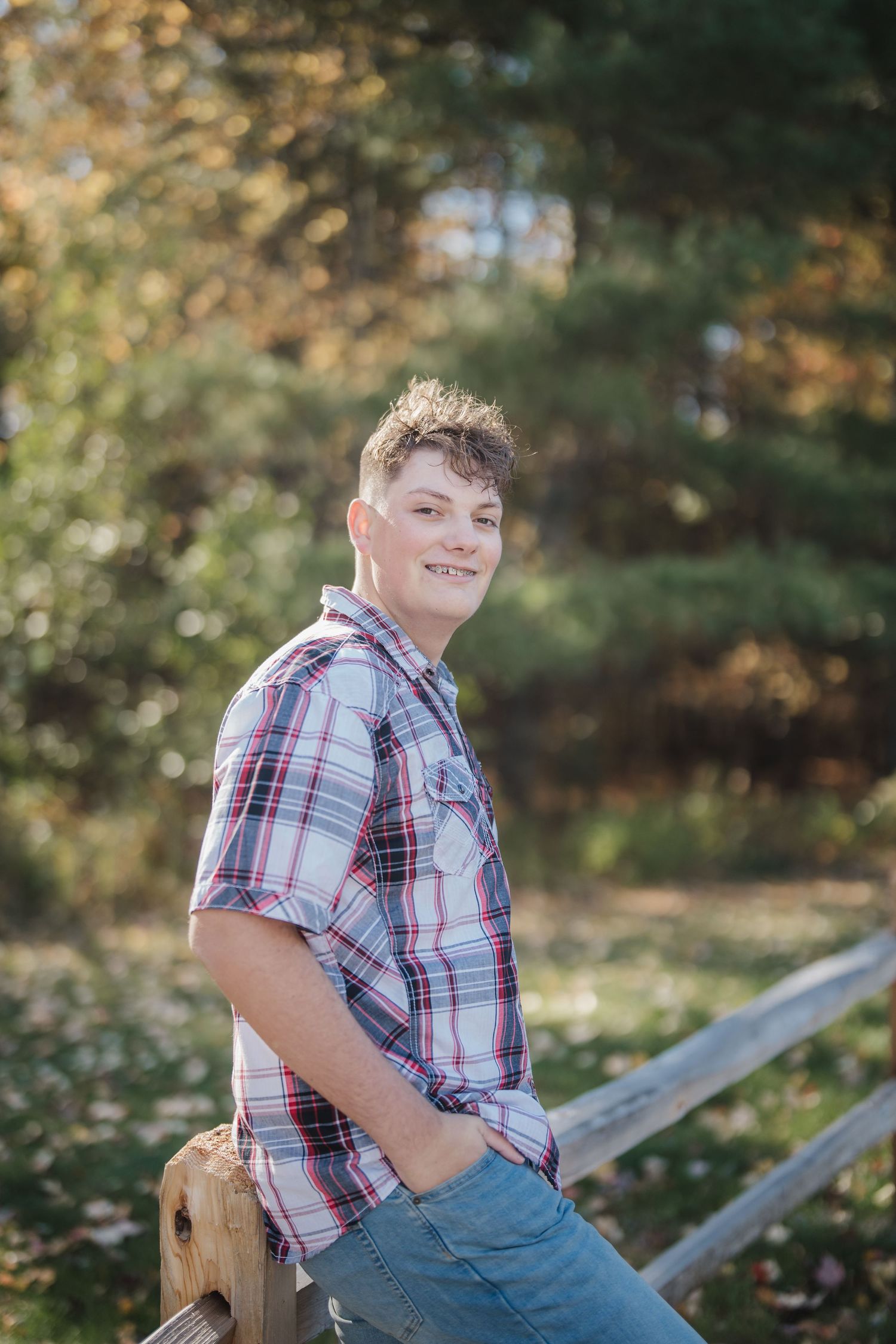Young person in plaid shirt and jeans sits casually on wooden fence during autumn outdoor photo session.