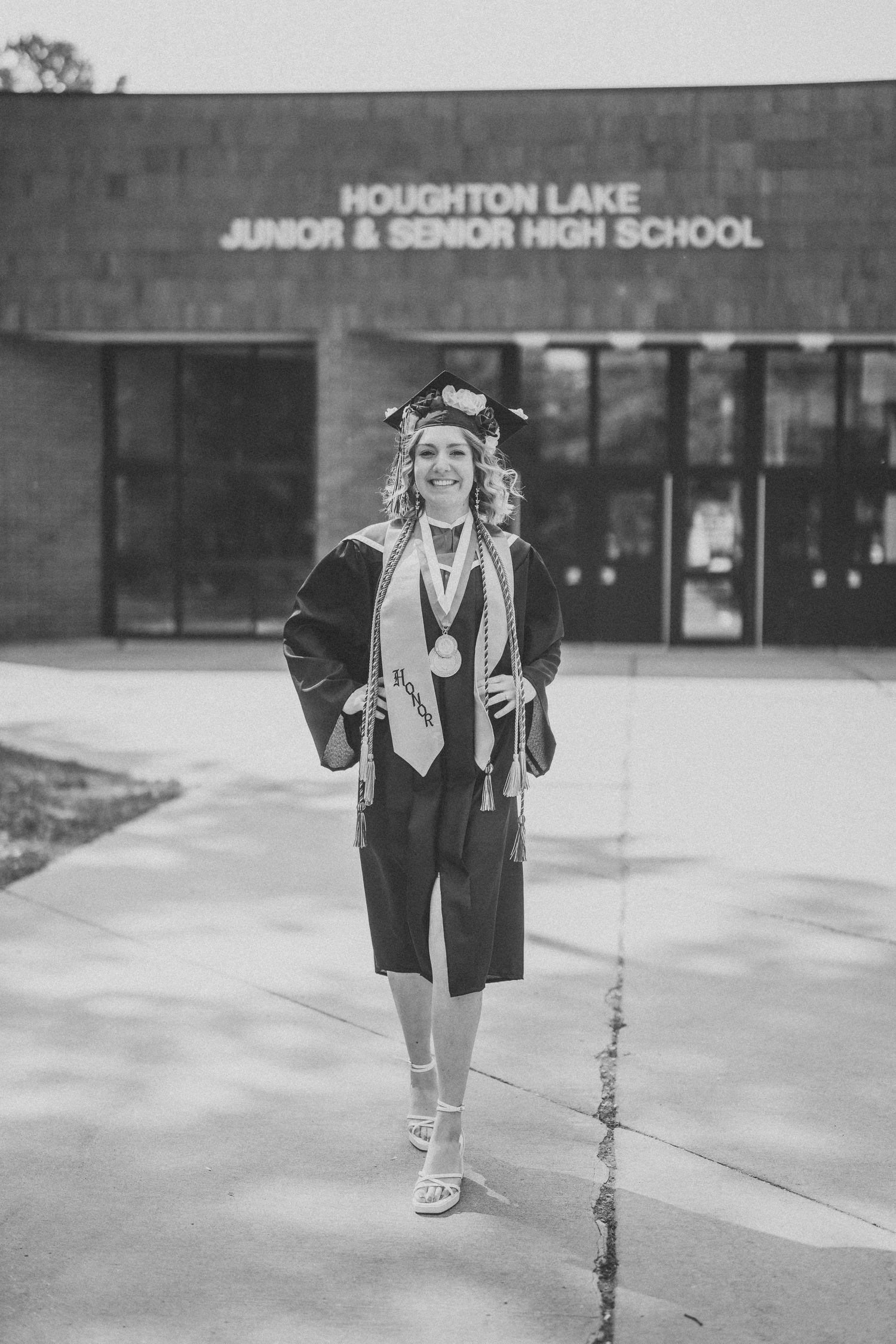A graduate poses in front of Houghton Lake Junior & Senior High School in a black graduation gown and cap.