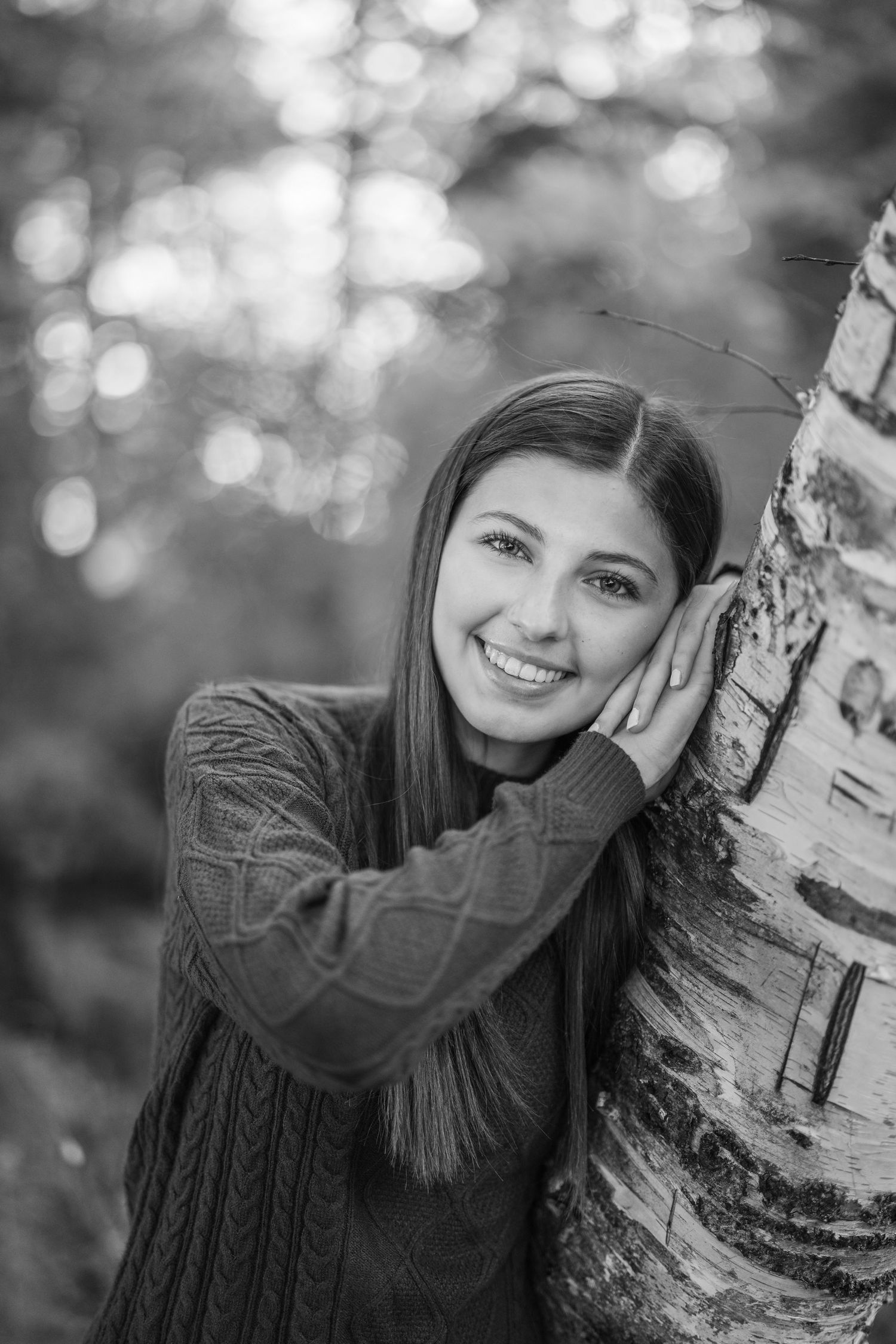 A black and white portrait of someone in a cable knit sweater leaning against a birch tree with bokeh background.
