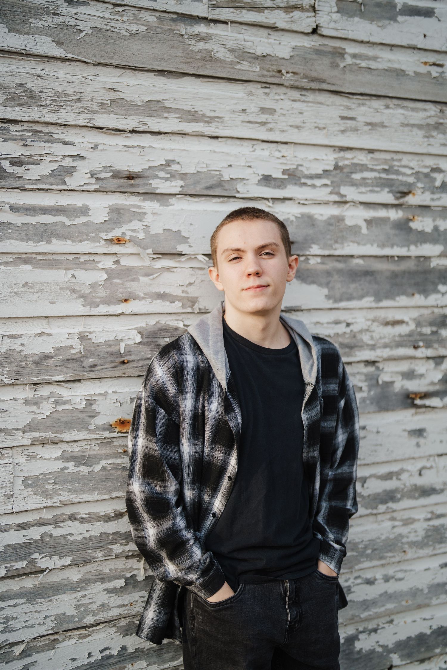 A casual portrait against weathered white wooden siding wearing a plaid flannel and dark clothing.