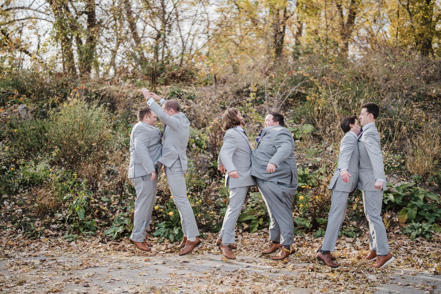 Groomsmen in light gray suits pose playfully in an autumn outdoor setting.