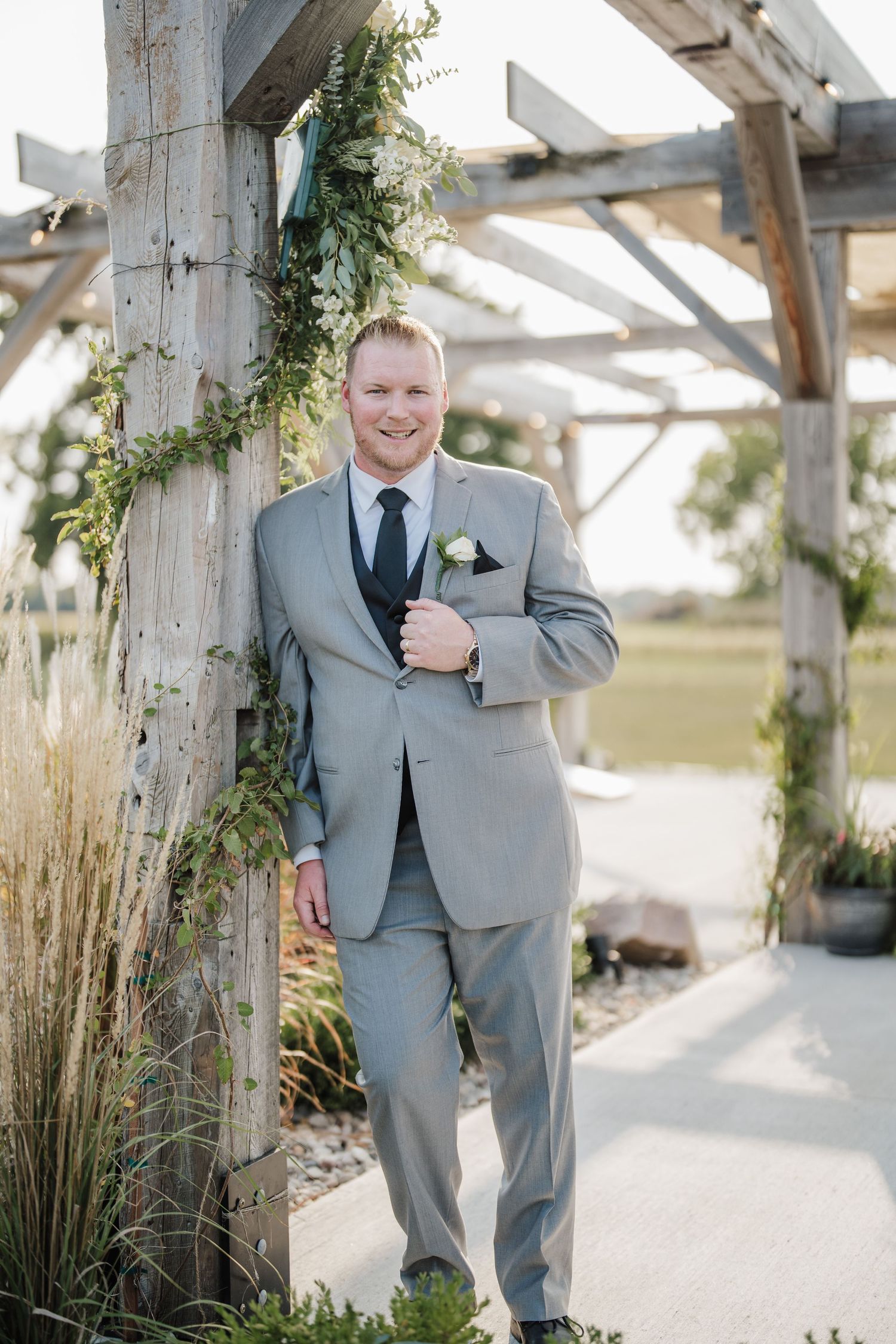A groom in grey suit smiles while standing under a decorated pergola.