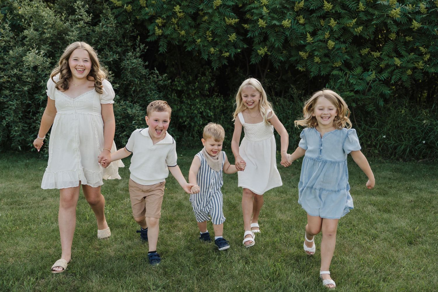 Five children holding hands and running together on green grass in front of lush bushes, wearing coordinated summer outfits.