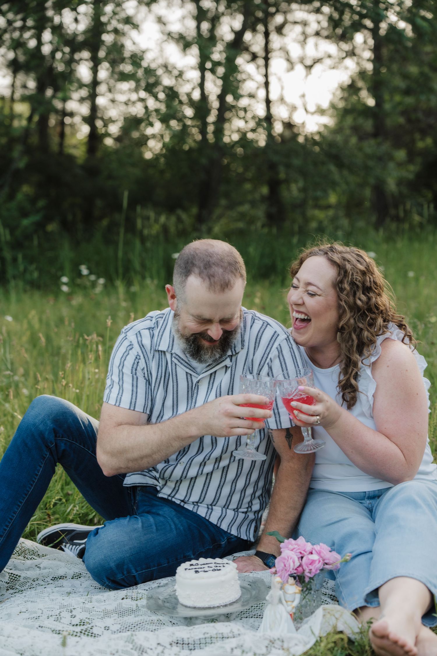 Couple laughing and toasting wine glasses during a romantic outdoor picnic on a blanket in a lush green field.