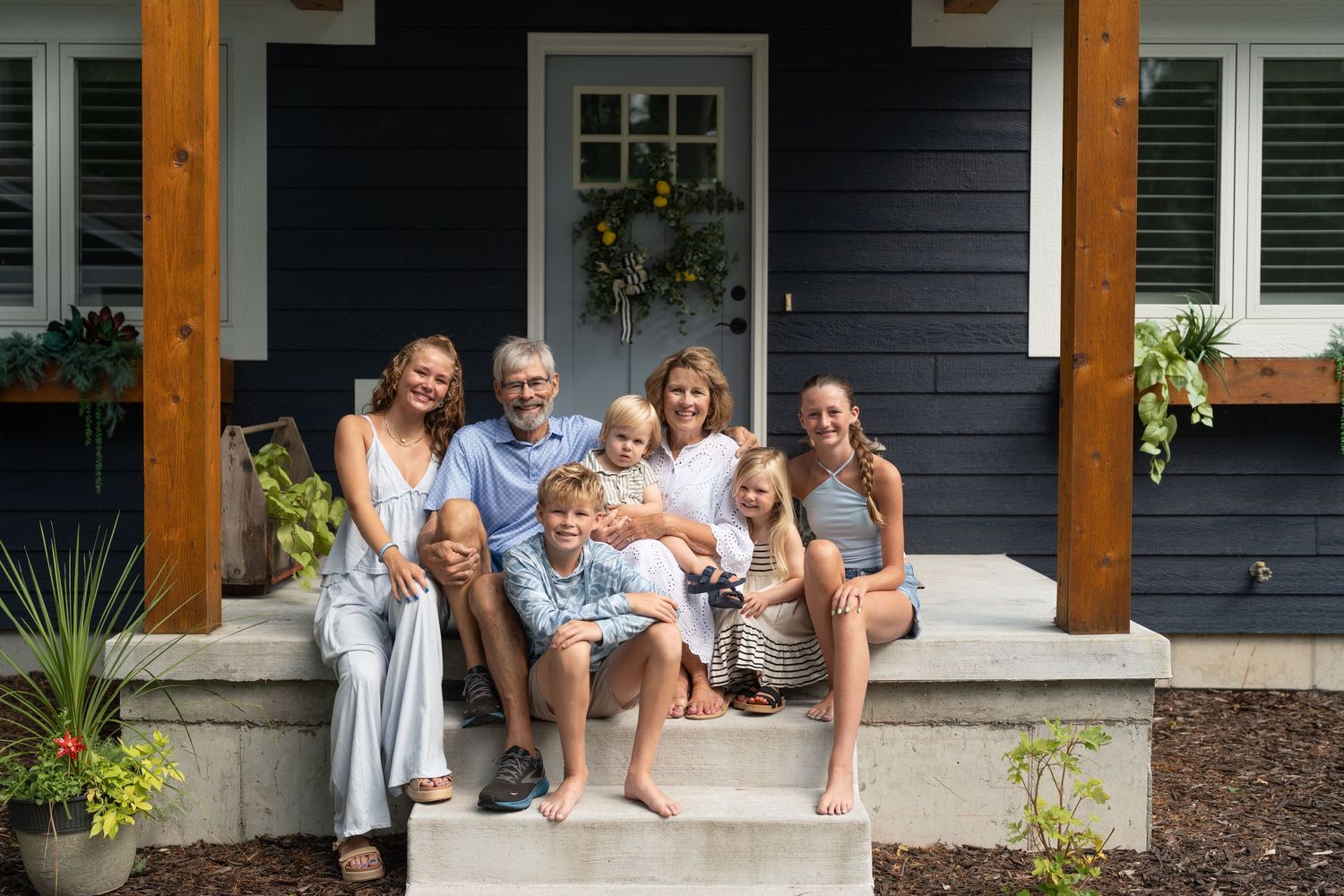 Multi-generational family smiling together on the front porch steps of a dark blue craftsman home.