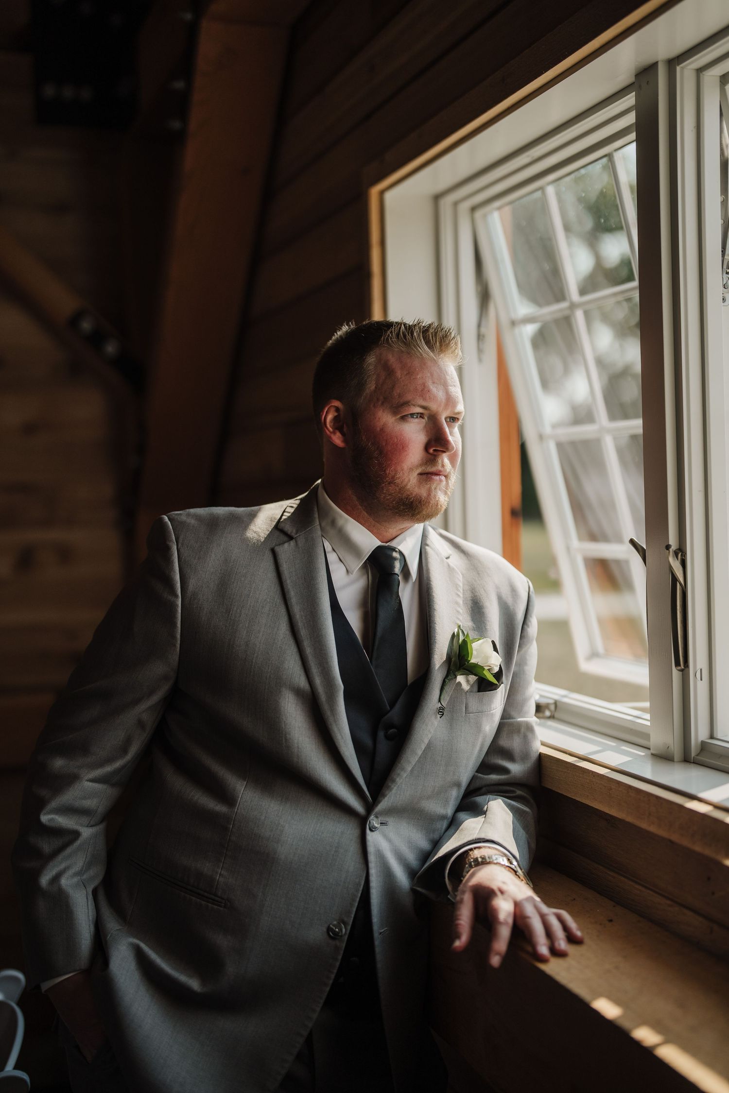 A groom in grey suit gazes thoughtfully out a rustic cabin window.