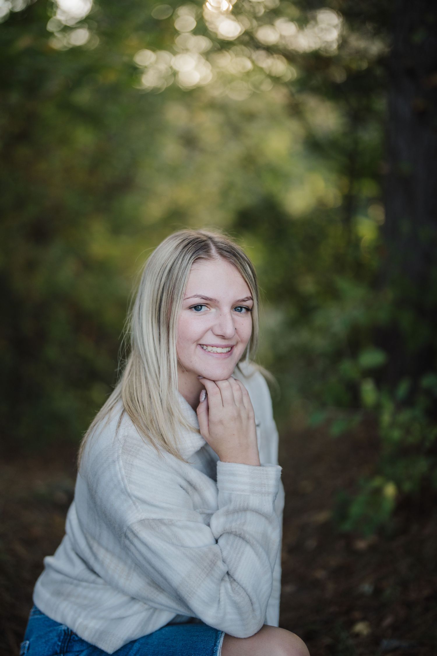 A person wearing a cozy gray sweater poses outdoors in a natural wooded setting with soft bokeh lighting effects.