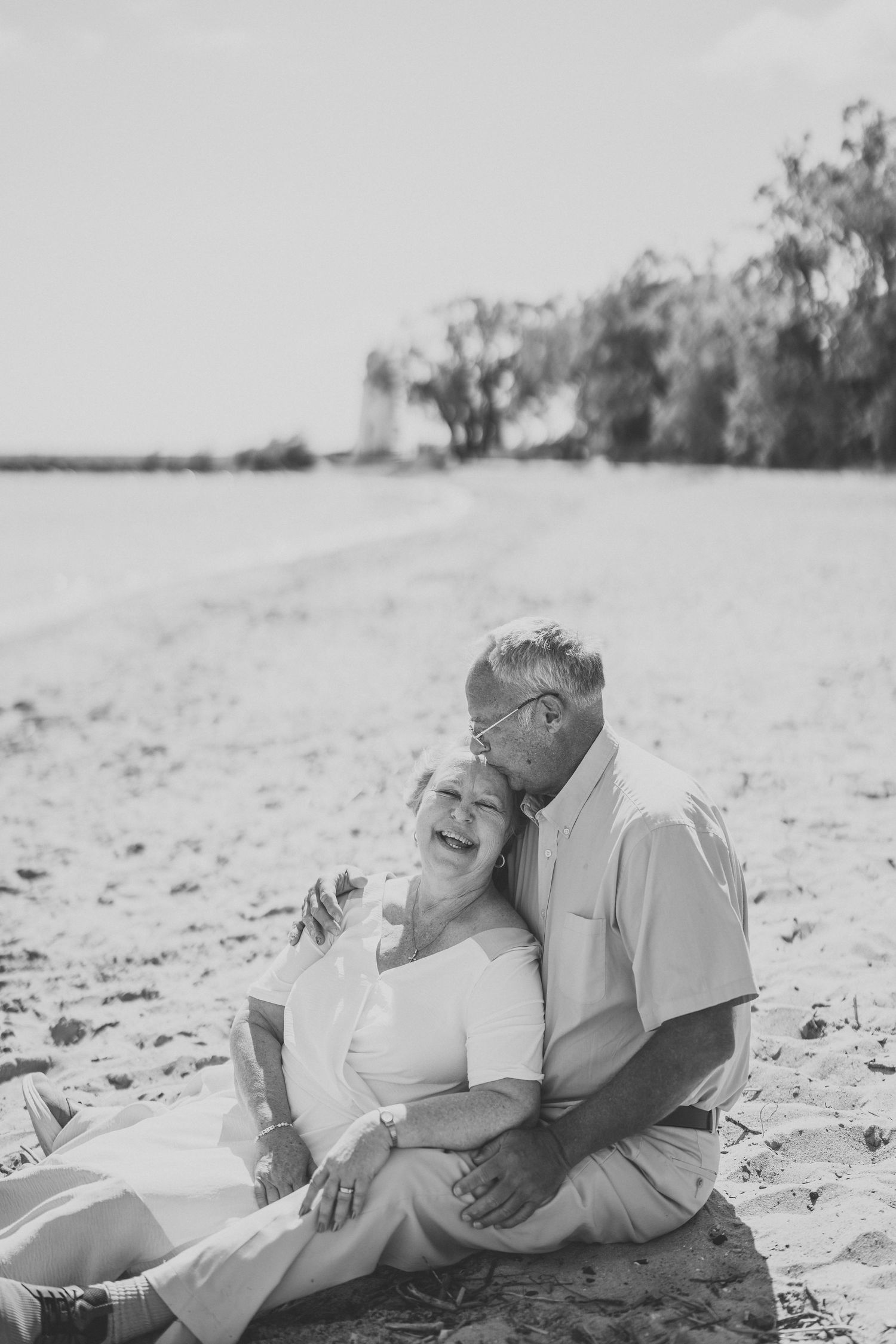 A black and white photo shows a couple lying together on a sandy beach in casual attire.