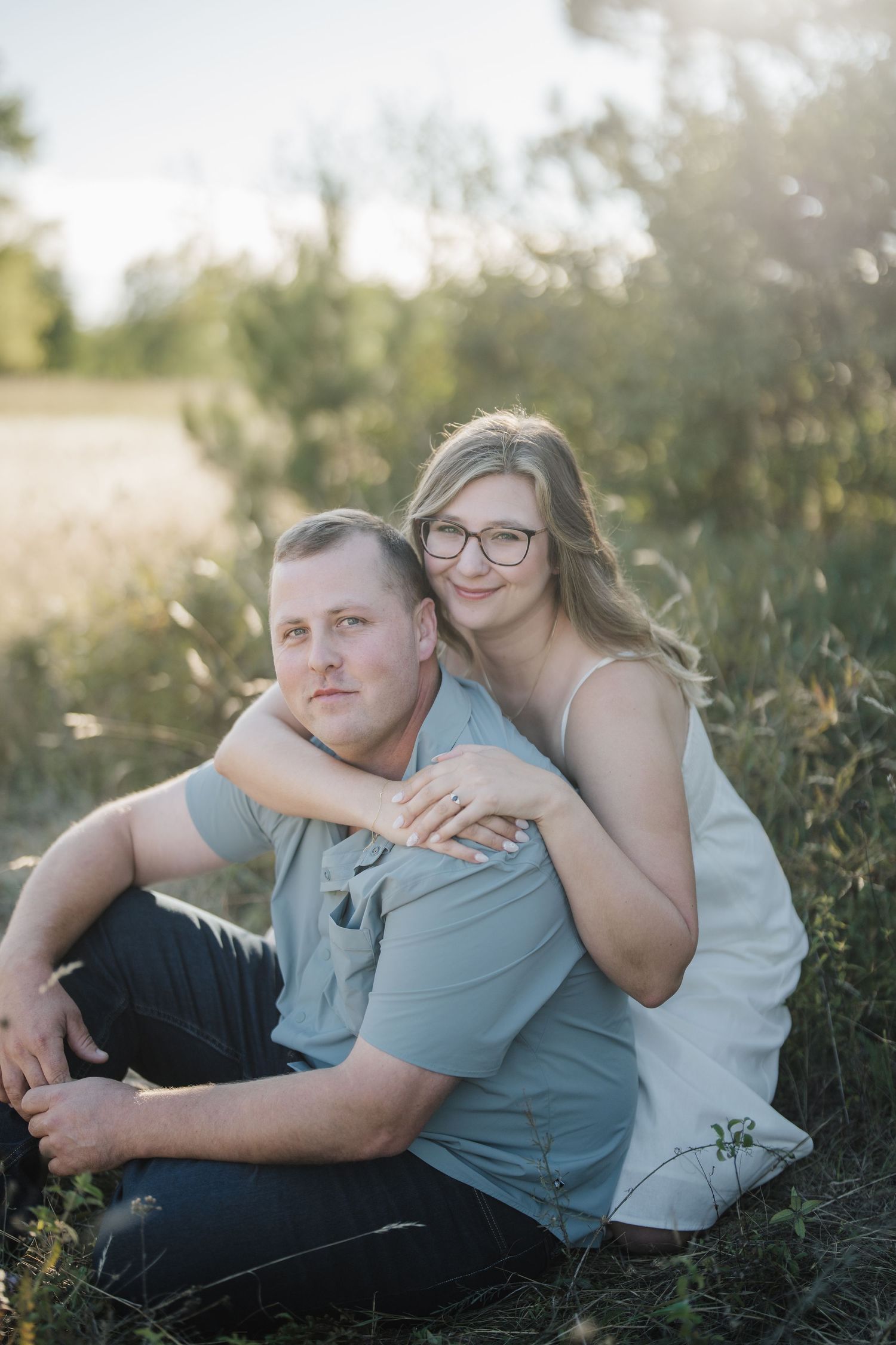 A couple sits together in a grassy field during golden hour, sharing an intimate moment in soft sunlight.