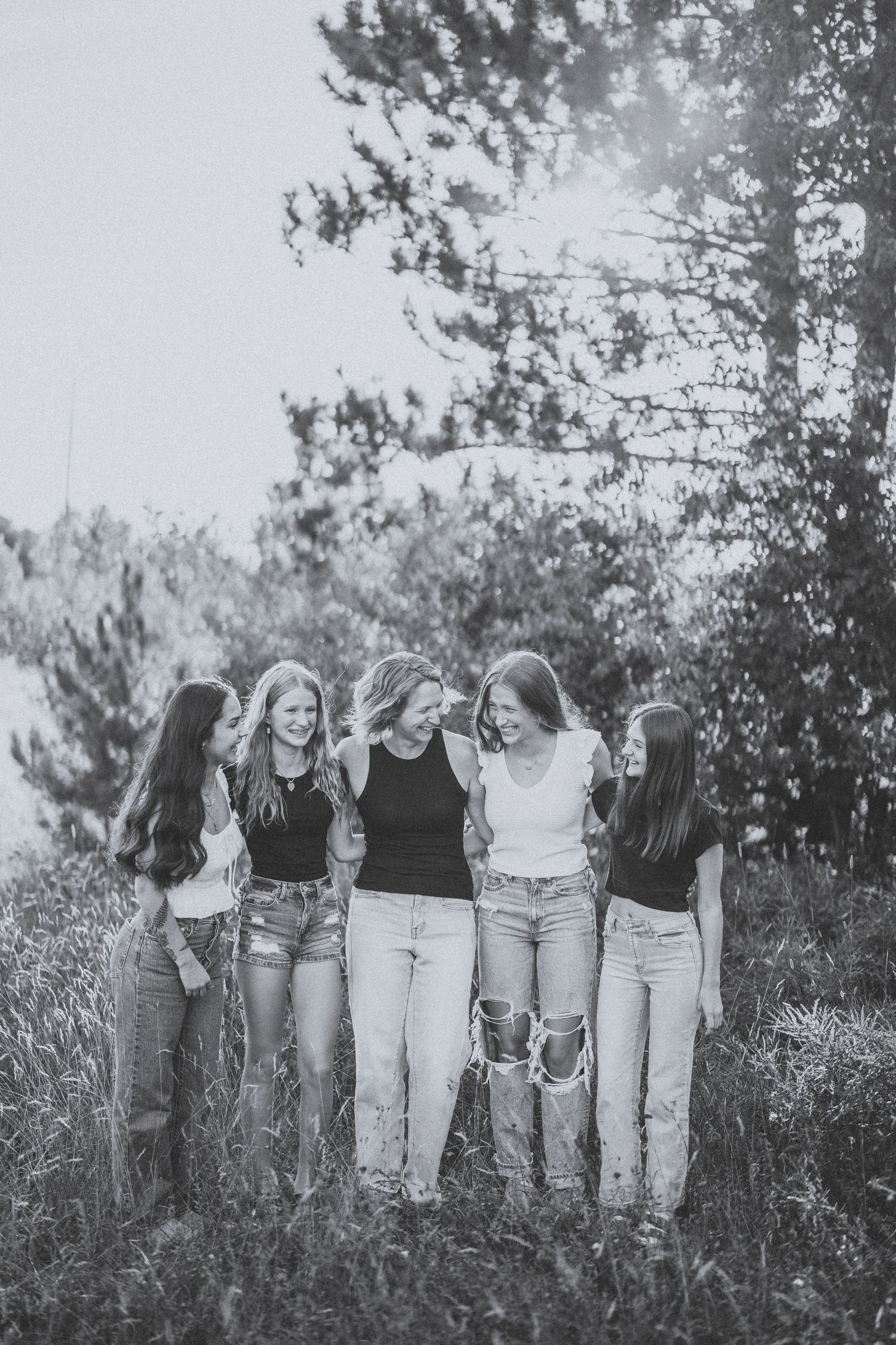 A group of friends in matching black tops and light jeans laugh together while standing in a grassy outdoor setting.