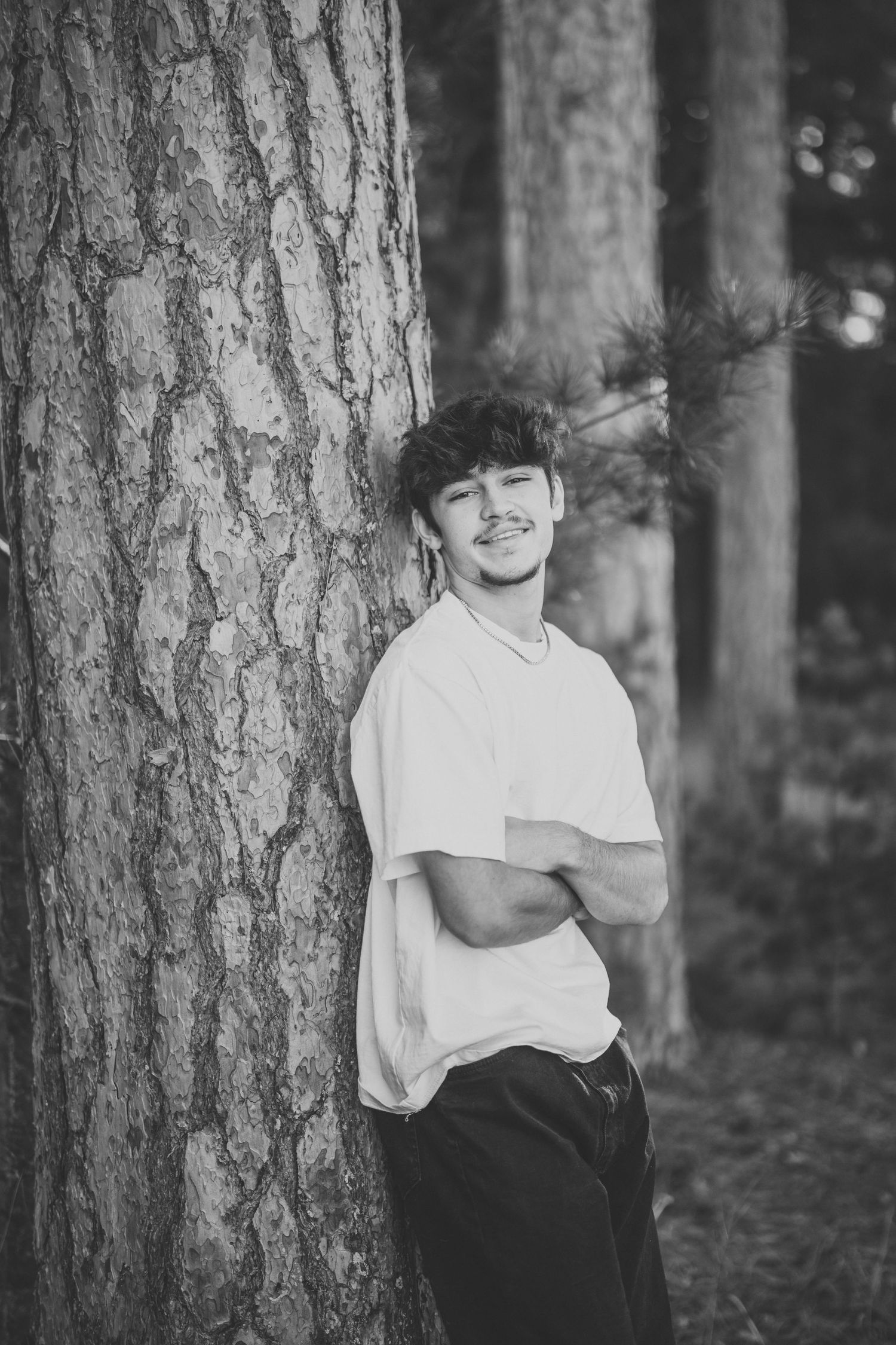 Black and white portrait of person leaning against large tree trunk in forest setting.