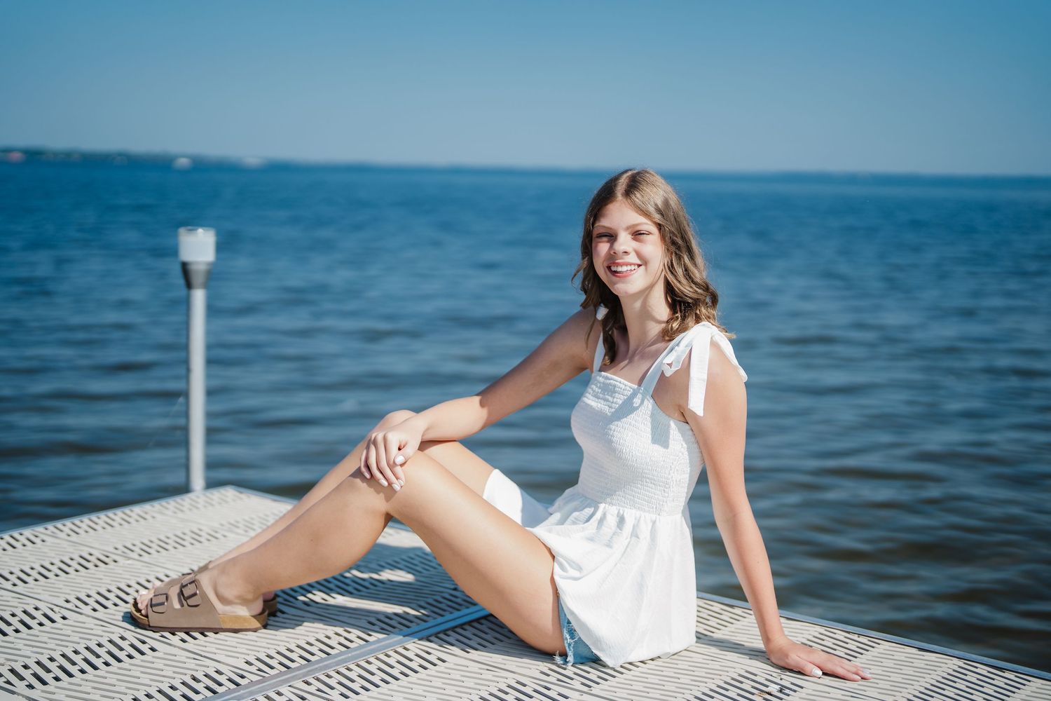 A person in a white summer dress sits on a dock by the blue ocean on a sunny day.