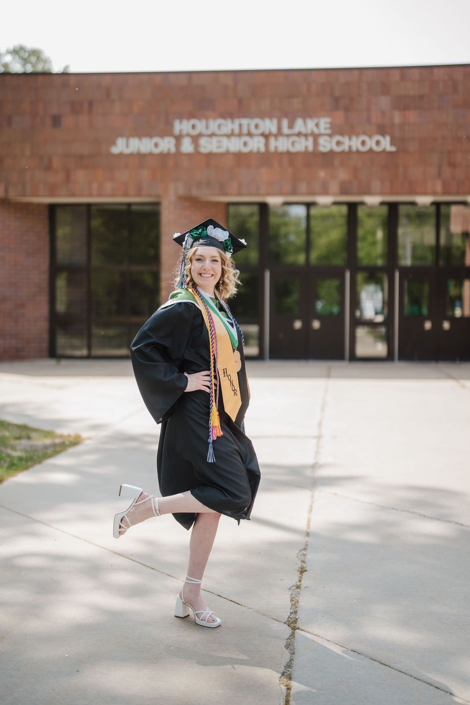 Graduate in cap and gown poses playfully outside Houghton Lake Junior & Senior High School entrance.