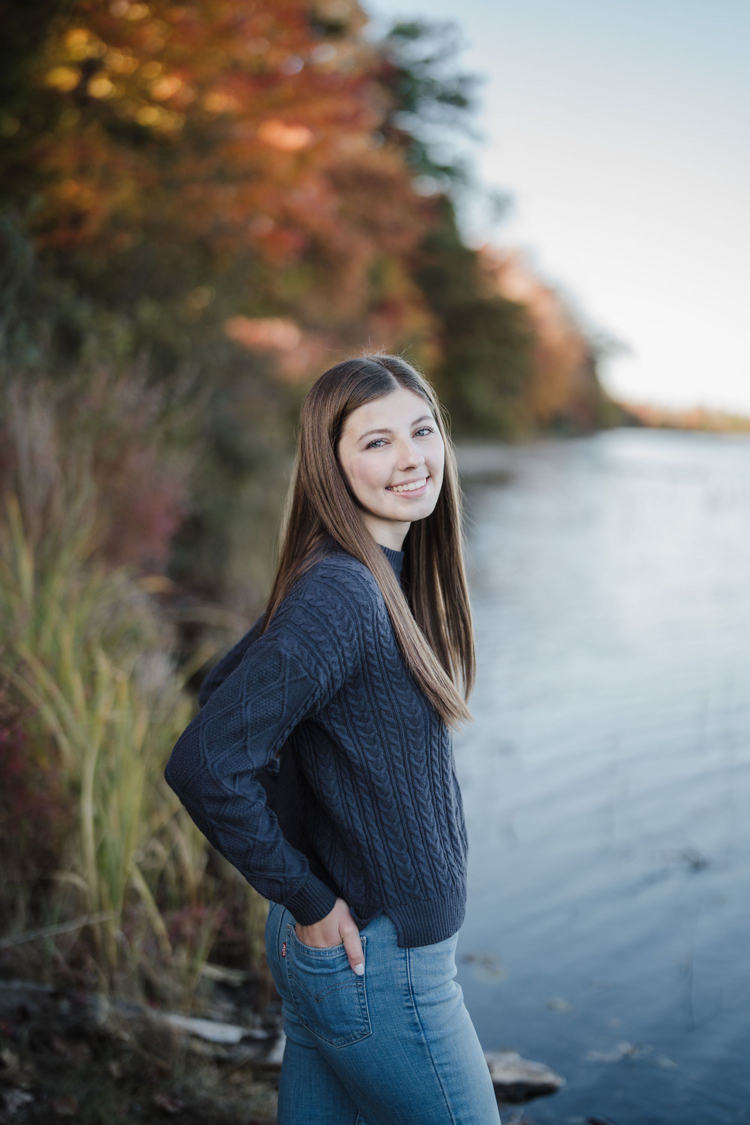 A person in a navy sweater stands by a waterfront with fall colors reflecting in the calm water.