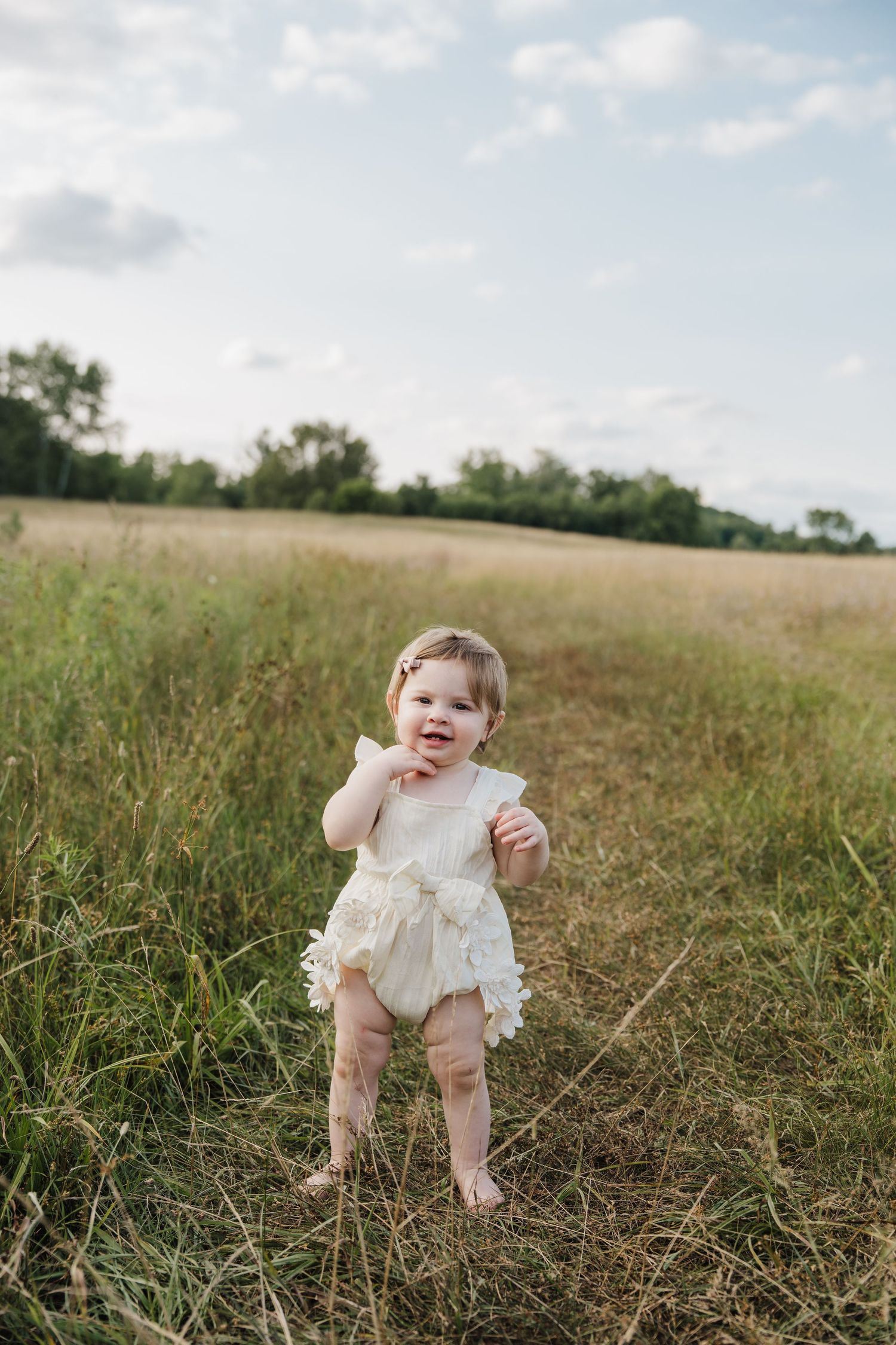 Adorable baby girl in white lace romper standing barefoot in a green meadow field under a blue sky.