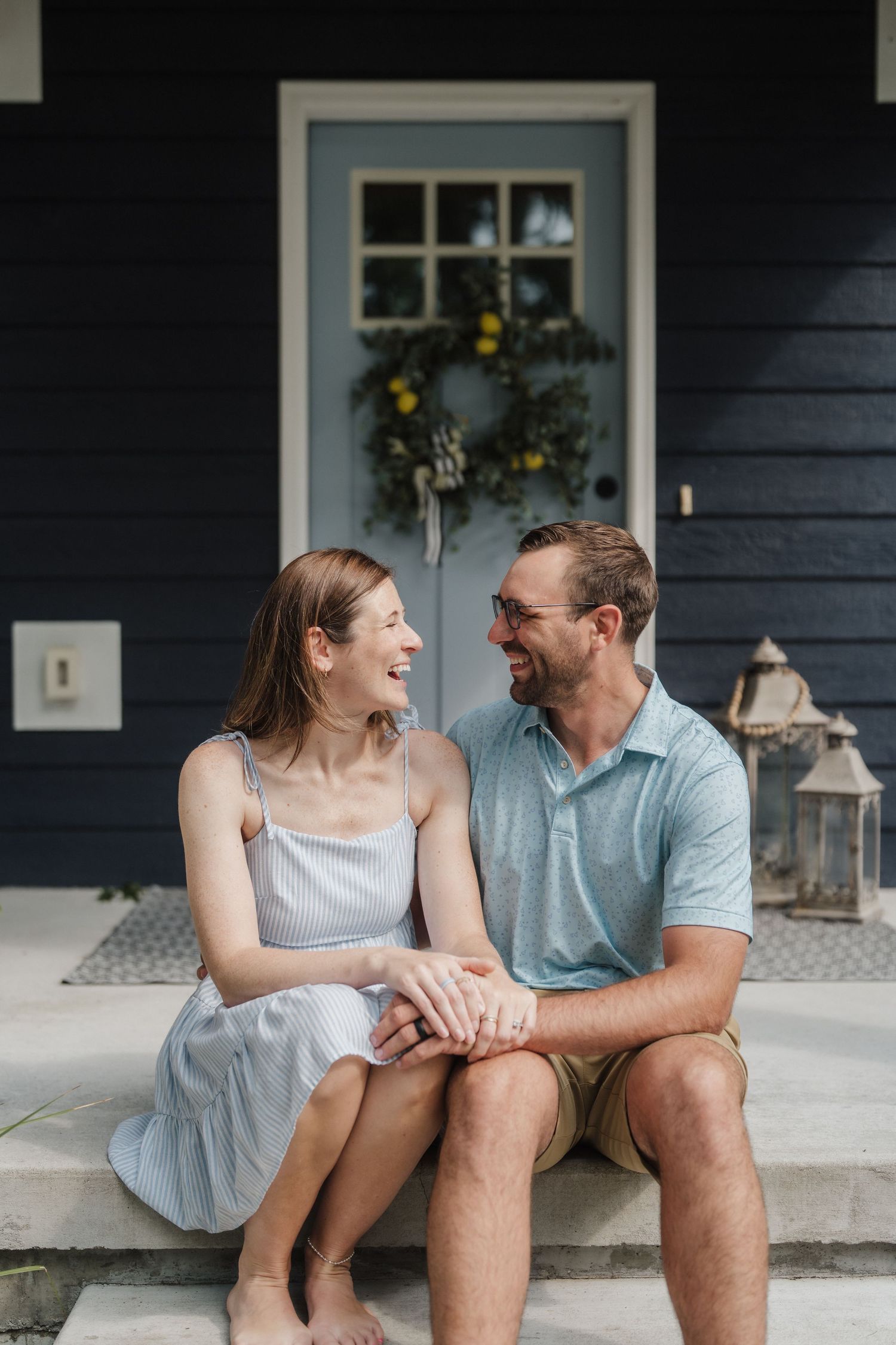 Happy couple sitting on front porch steps of a navy blue home with a lemon wreath on the door.