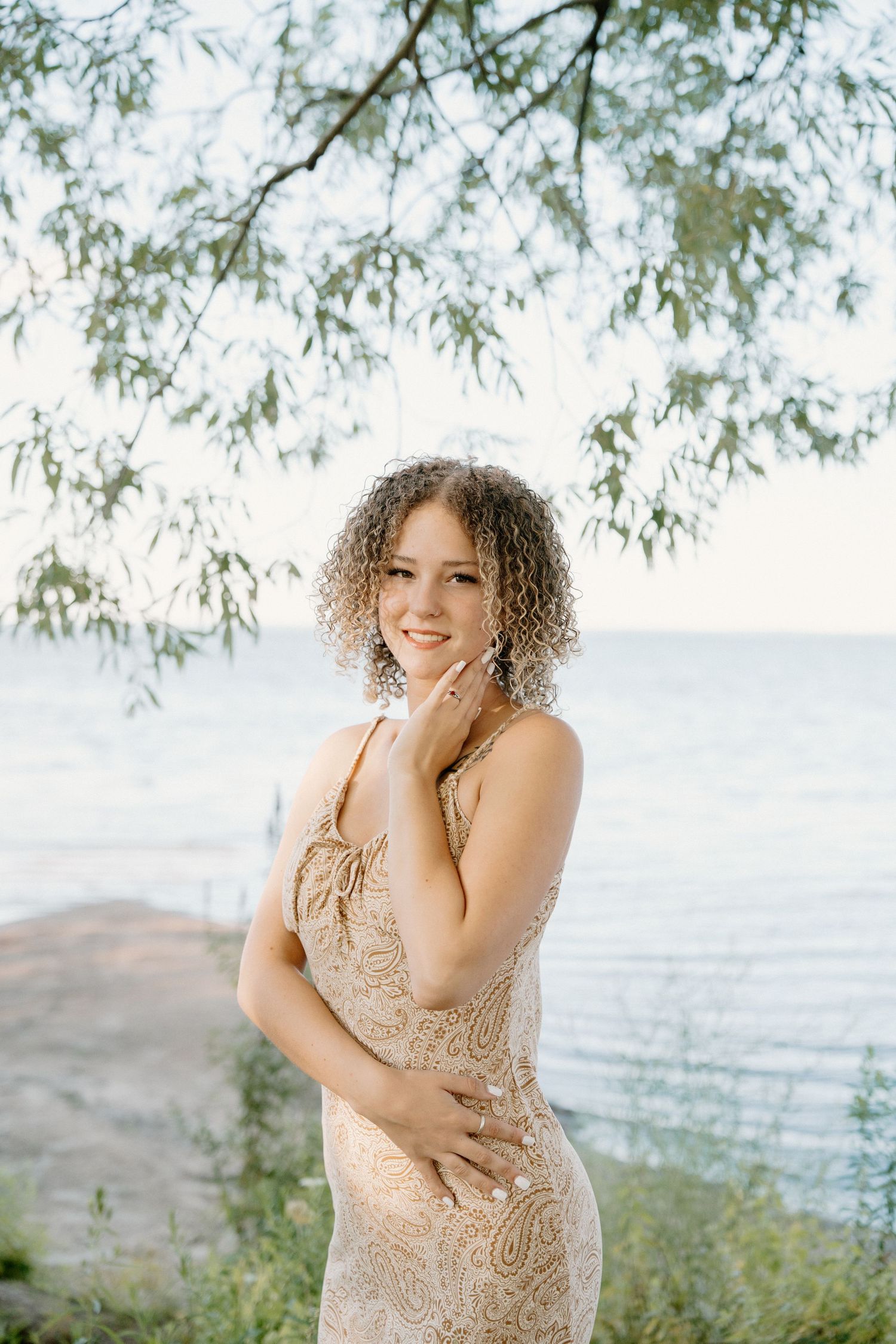 Young woman in gold lace dress smiling by the water under tree branches on a sunny day.