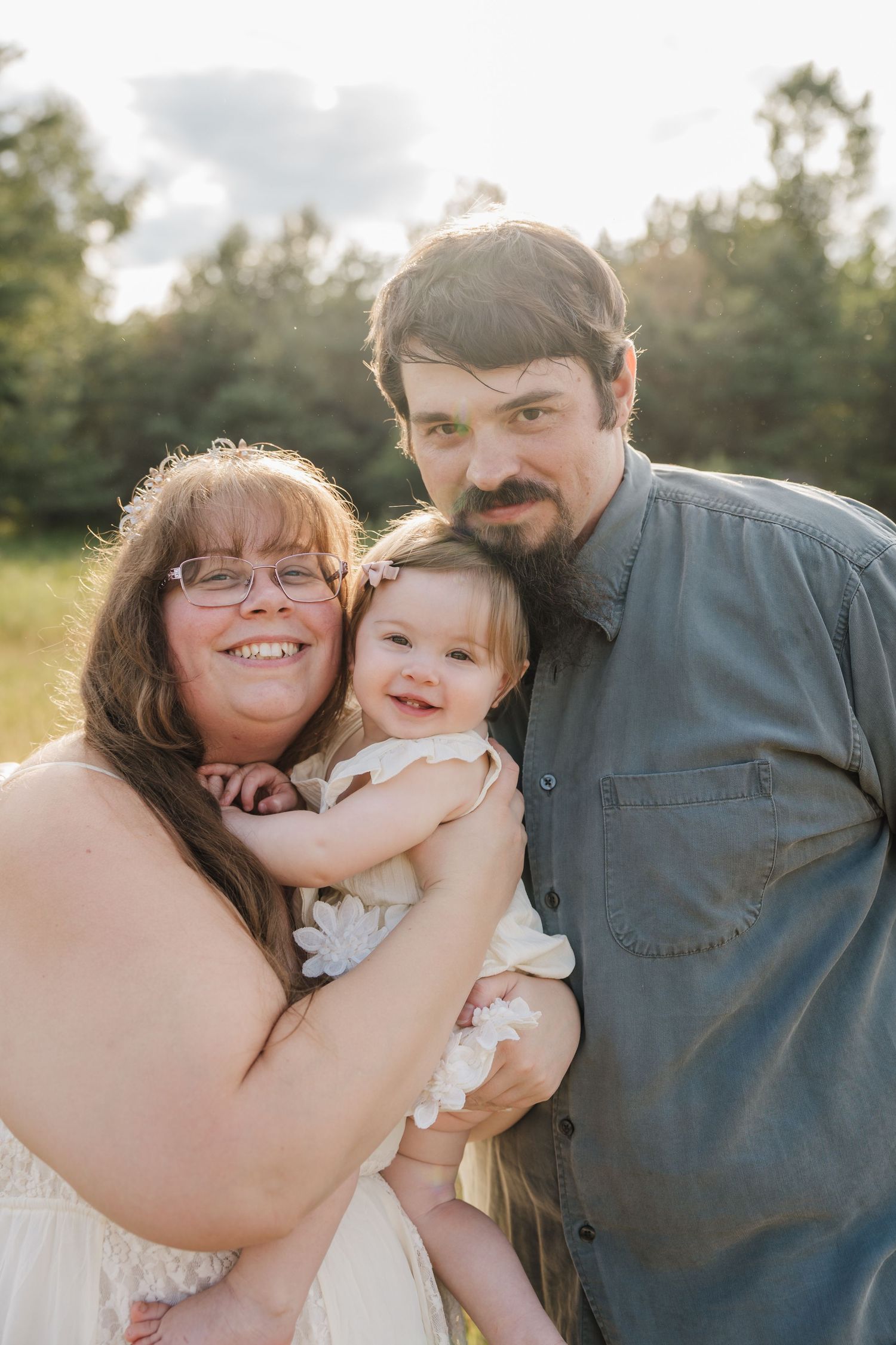 Happy family portrait of parents holding smiling baby girl outdoors in golden hour sunlight with green trees behind.