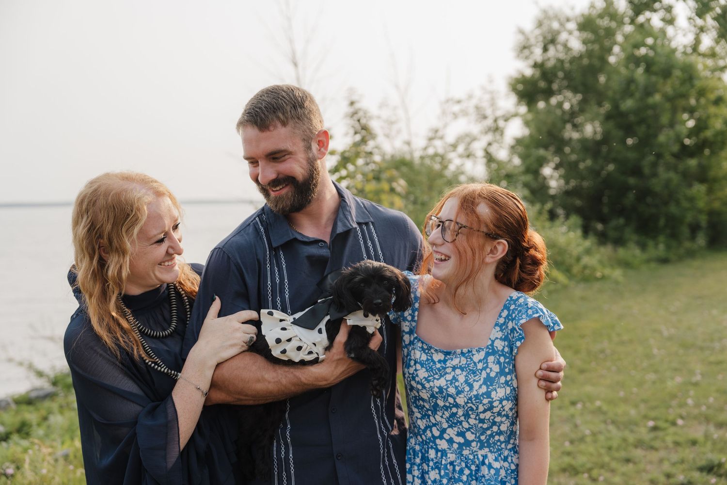 Two redhead women and a bearded man holding a small black puppy outdoors near a lake.