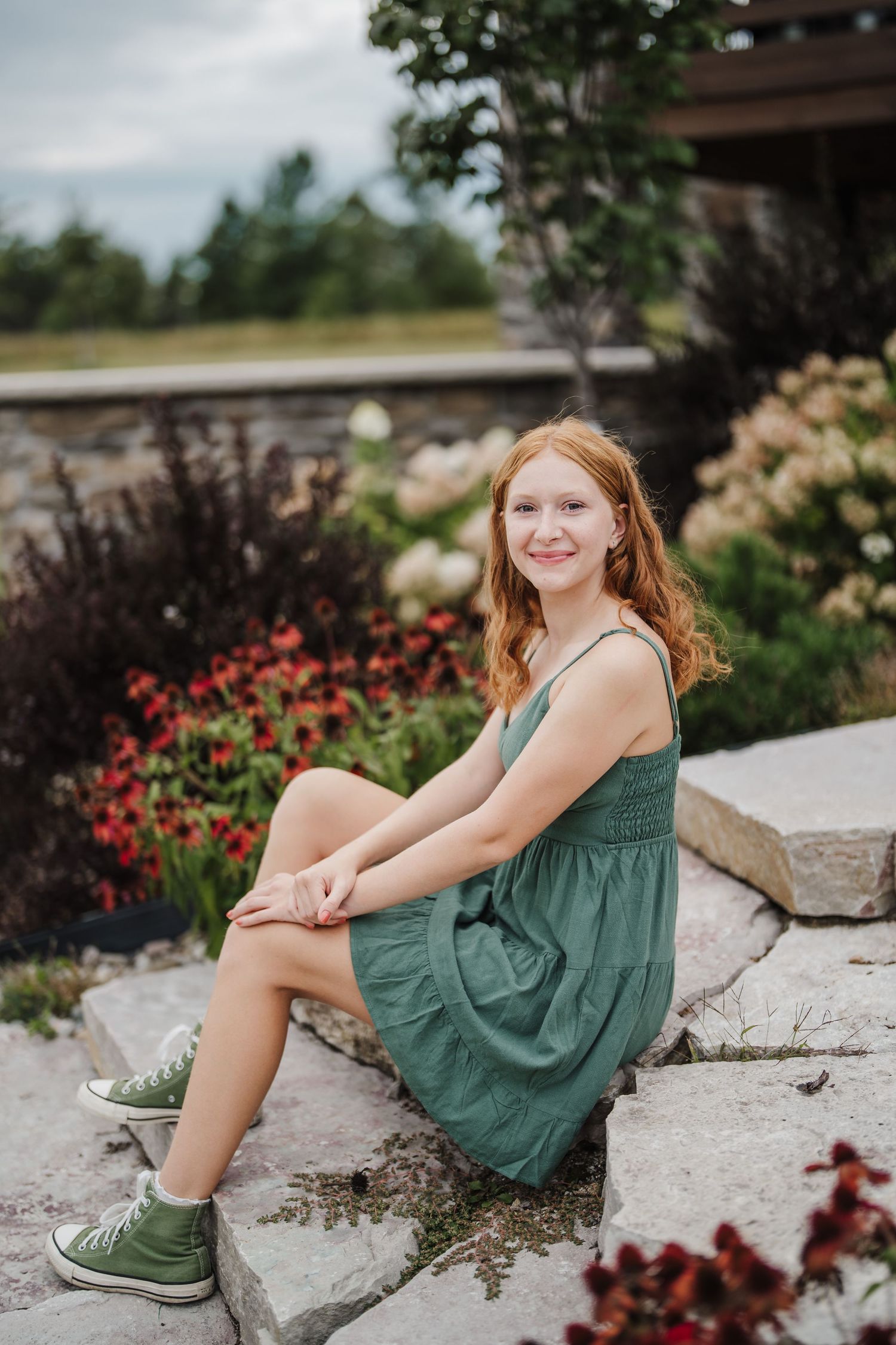 Person in green summer dress and sneakers sits on stone steps surrounded by colorful garden flowers.