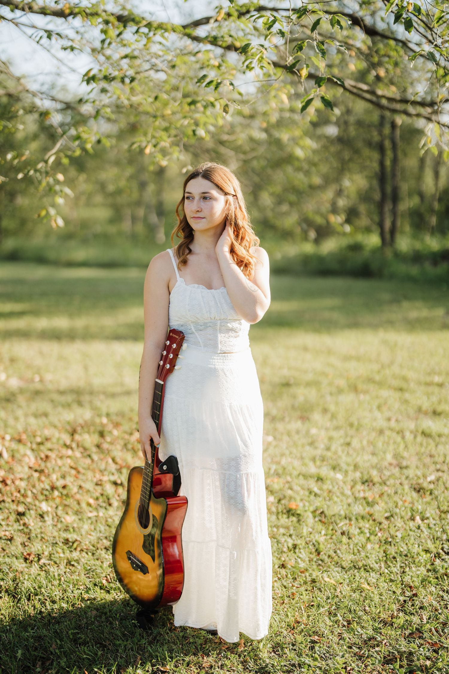 A musician in a white dress poses with an acoustic guitar in a sunny meadow with green foliage.