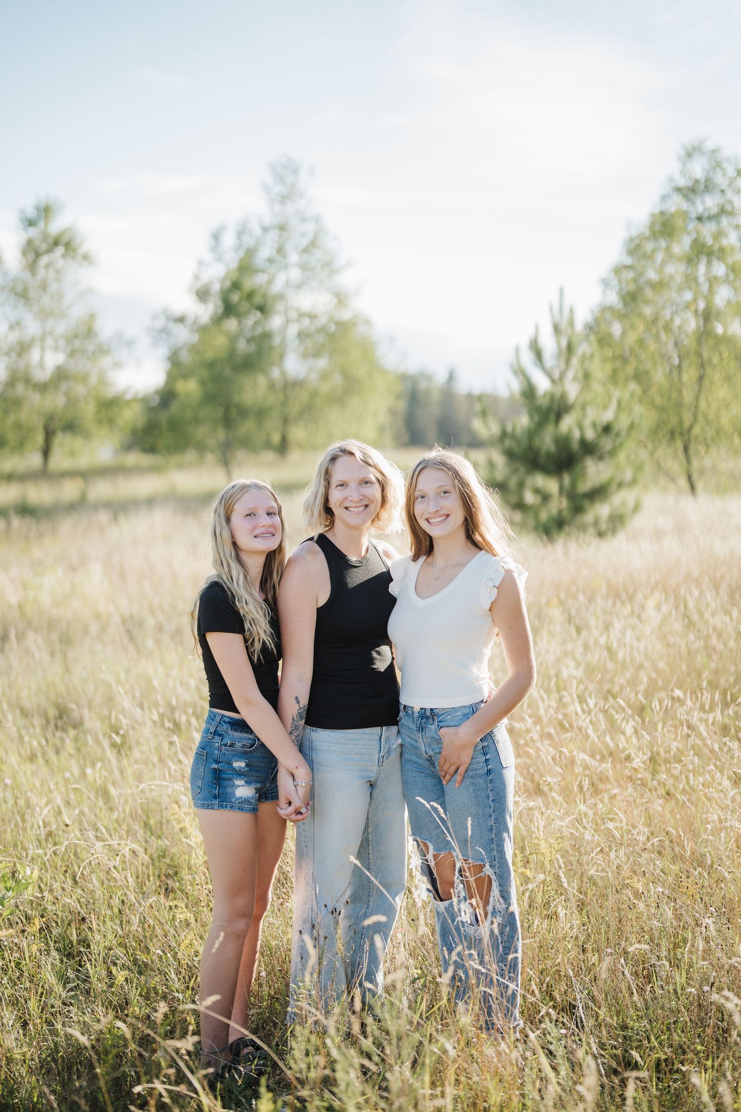 Three friends stand together in a sunlit meadow with tall grass and trees in the background on a bright summer day.