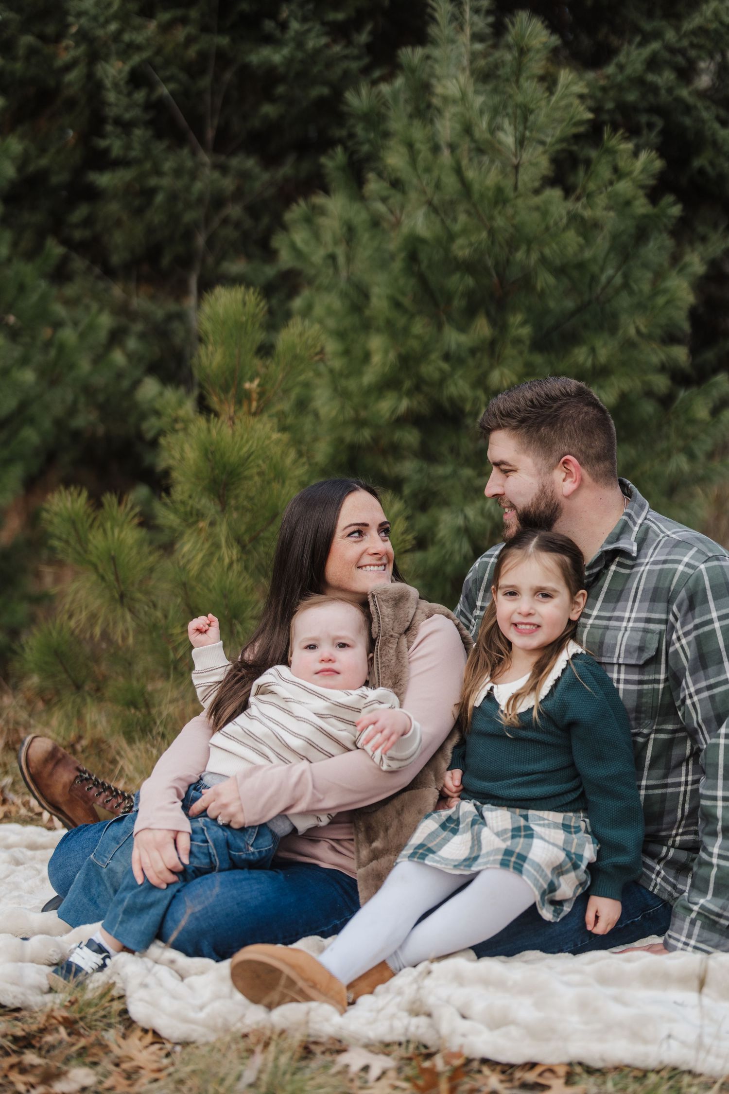Family sits together on white blanket in front of evergreen trees during outdoor portrait session.