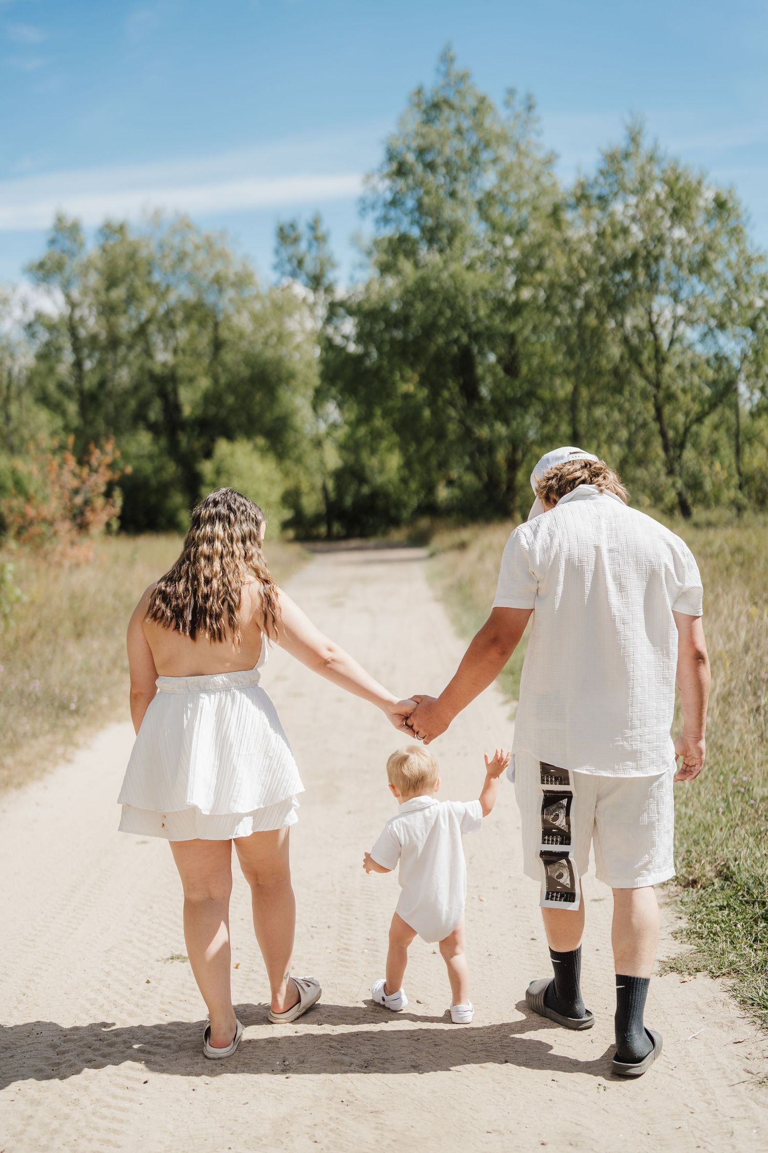 Family wearing white clothing walks together on a sandy path through nature, photographed from behind.