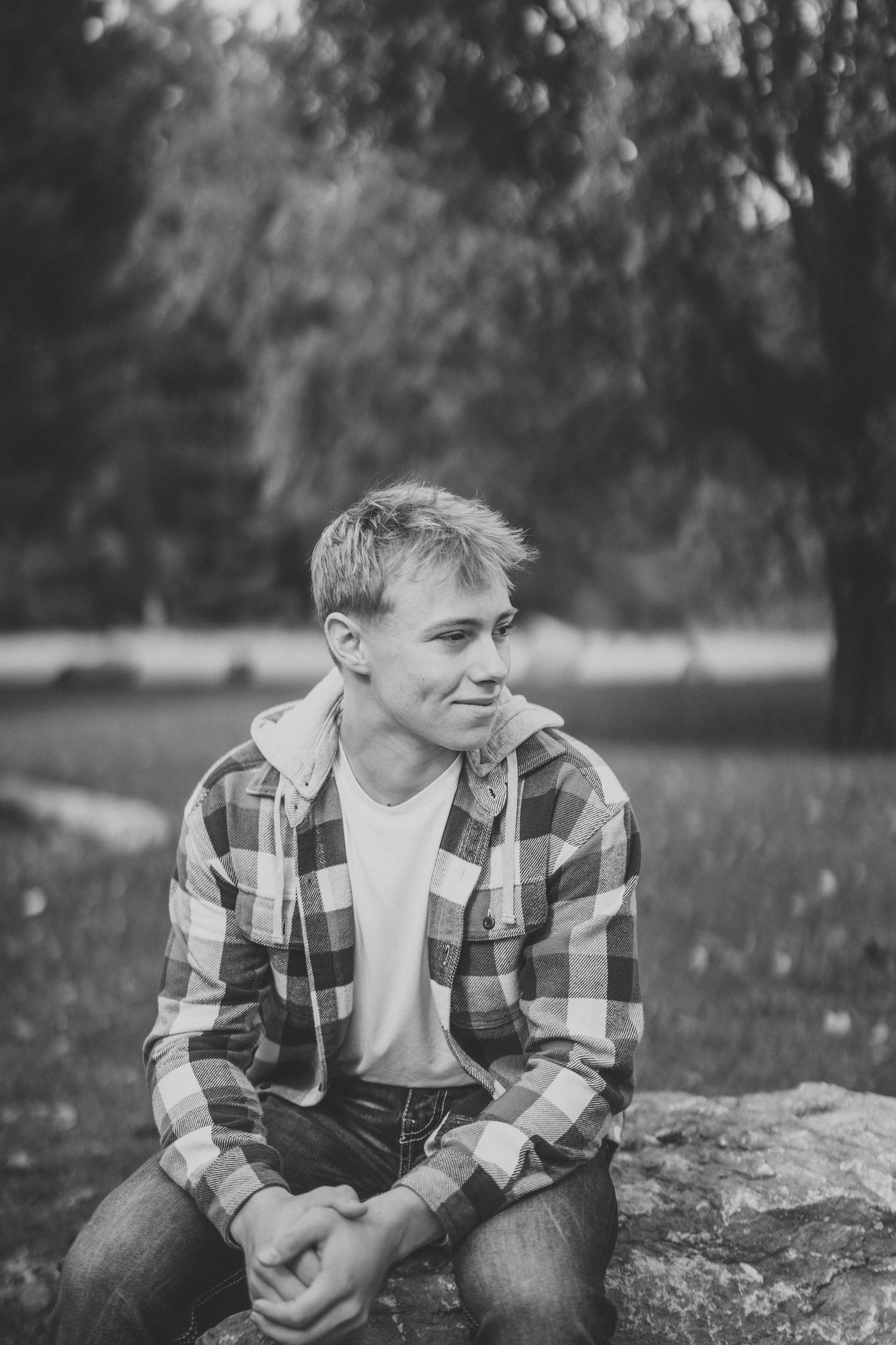 Black and white portrait of someone in a plaid shirt sitting on rocks outdoors while looking off to the side.
