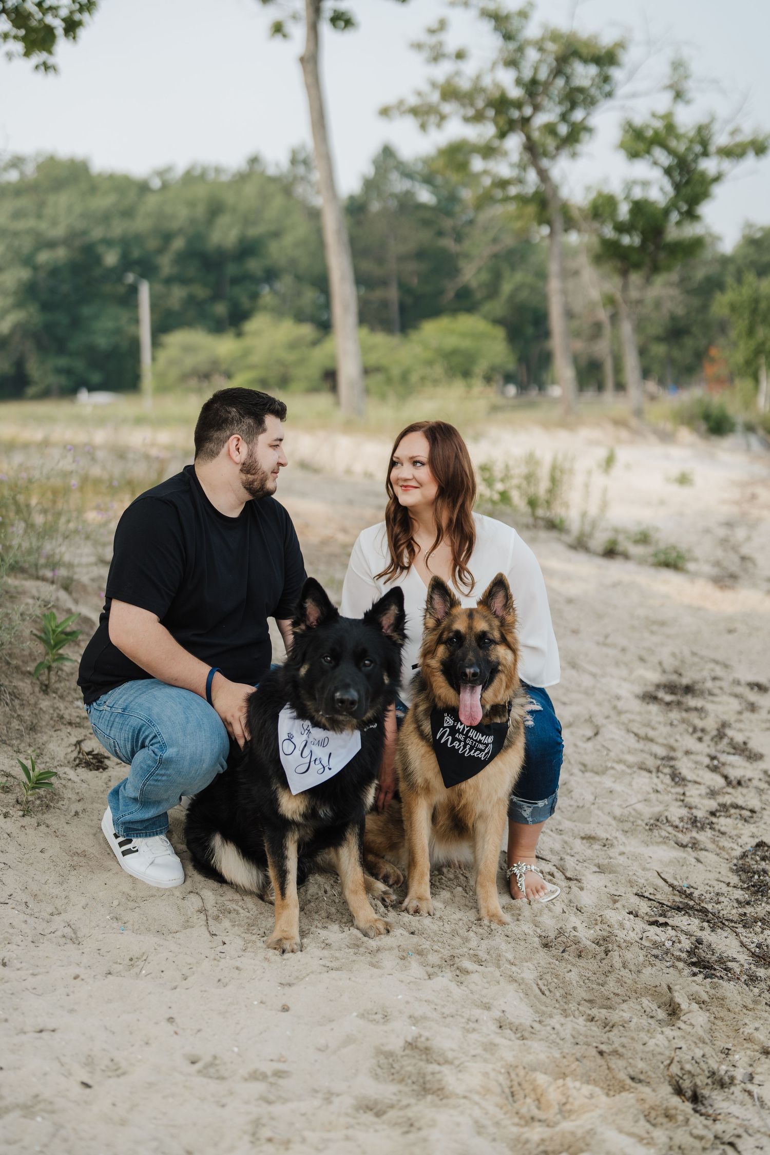 A couple sits on a sandy beach with their two German Shepherd dogs enjoying a relaxed outdoor moment together.