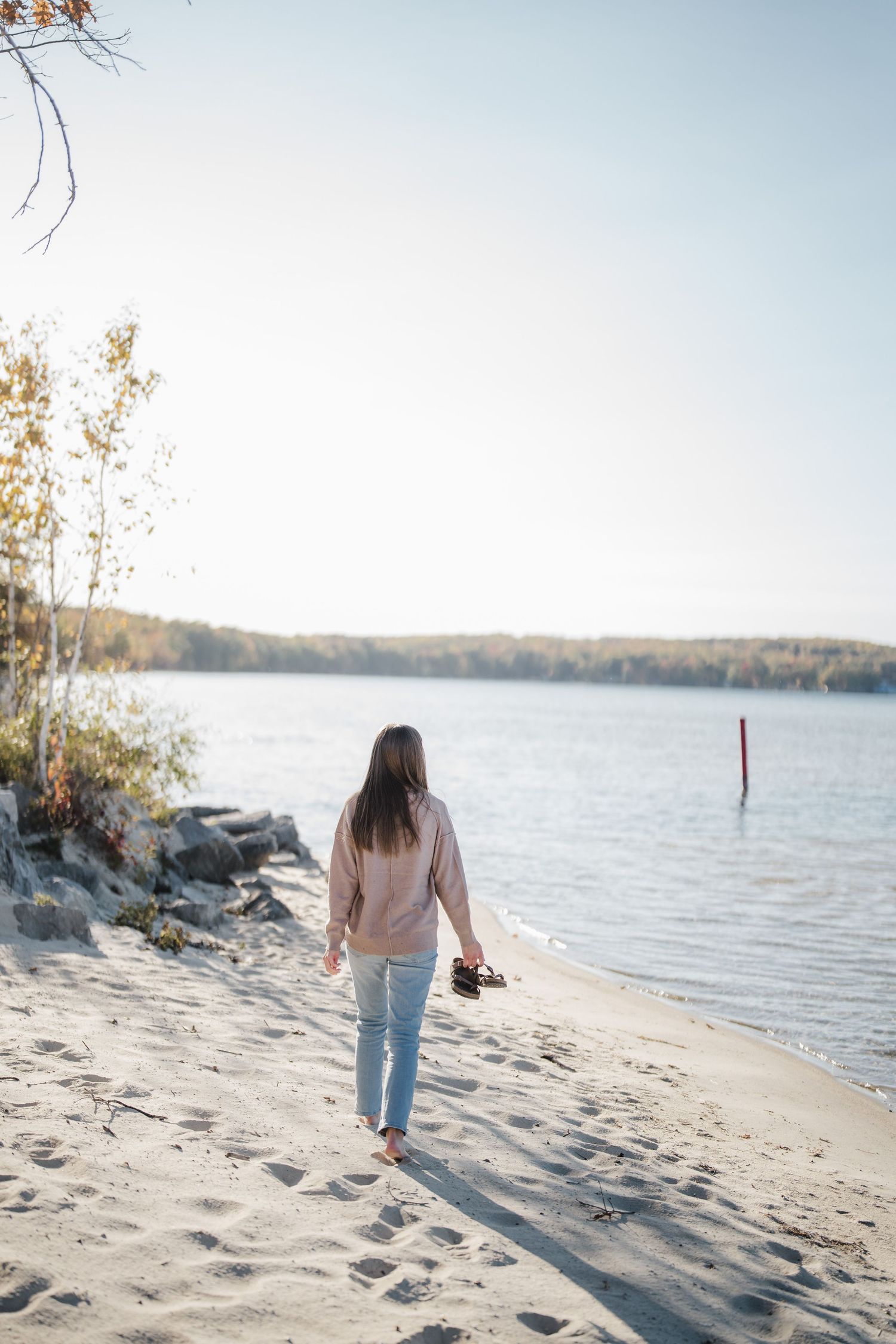 A person in jeans walks along a sandy beach shoreline on an autumn day with calm waters and distant trees.