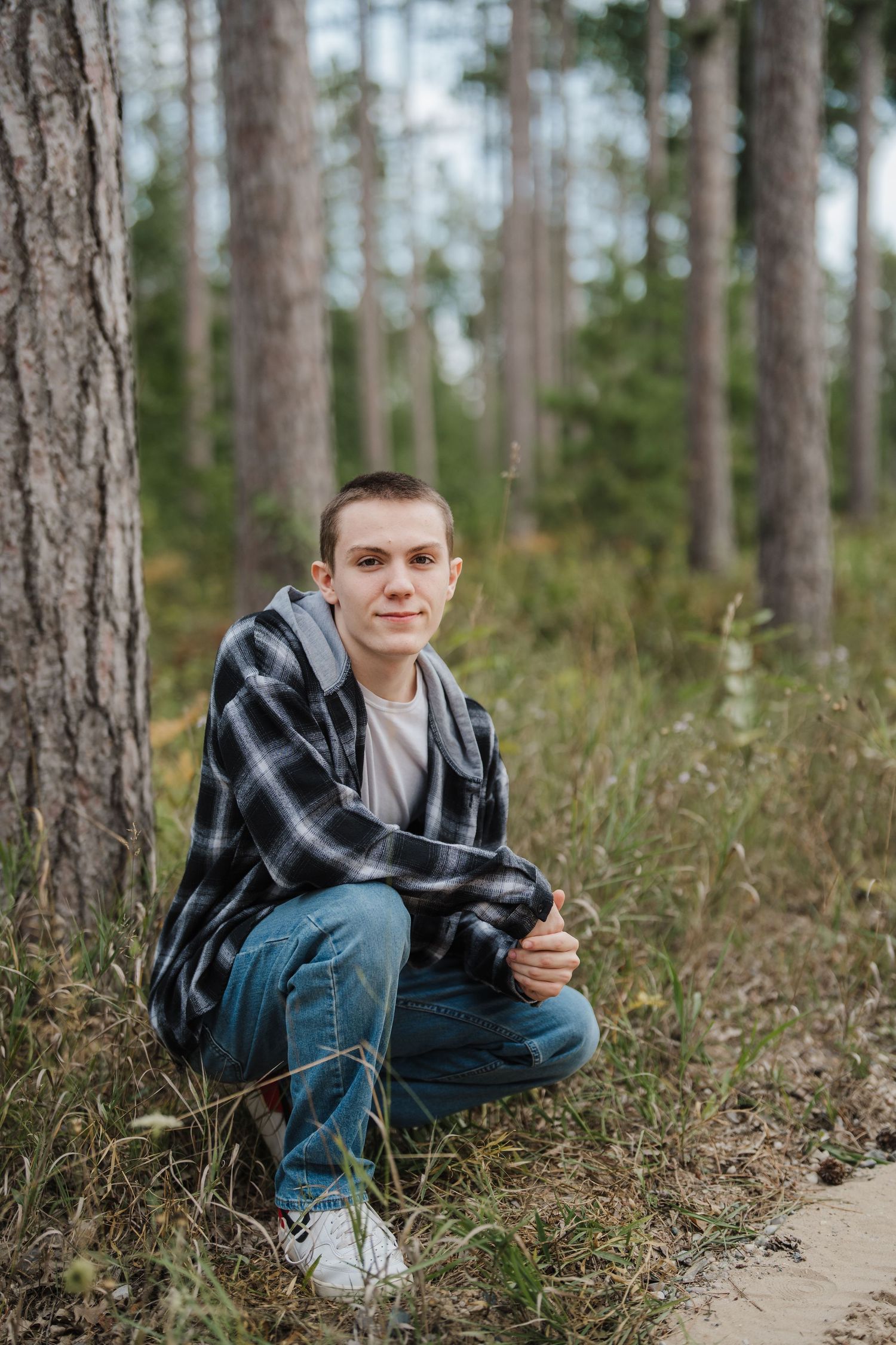 A person in a plaid shirt and jeans sits casually on a forest hillside among tall pine trees.