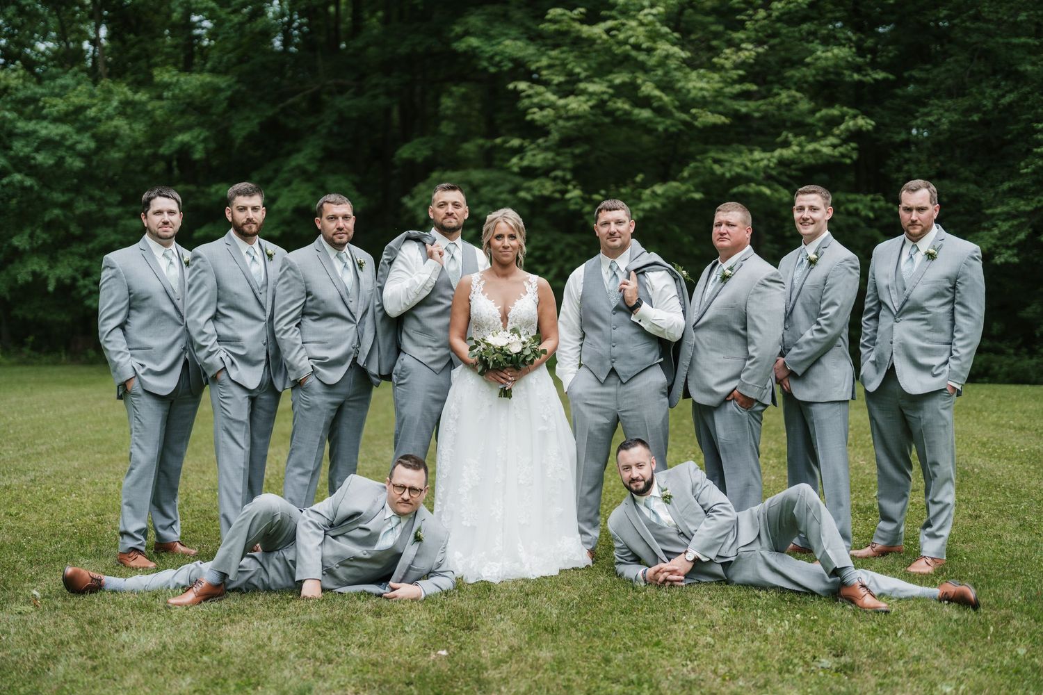 Wedding party poses playfully on lawn with groomsmen in light gray suits.