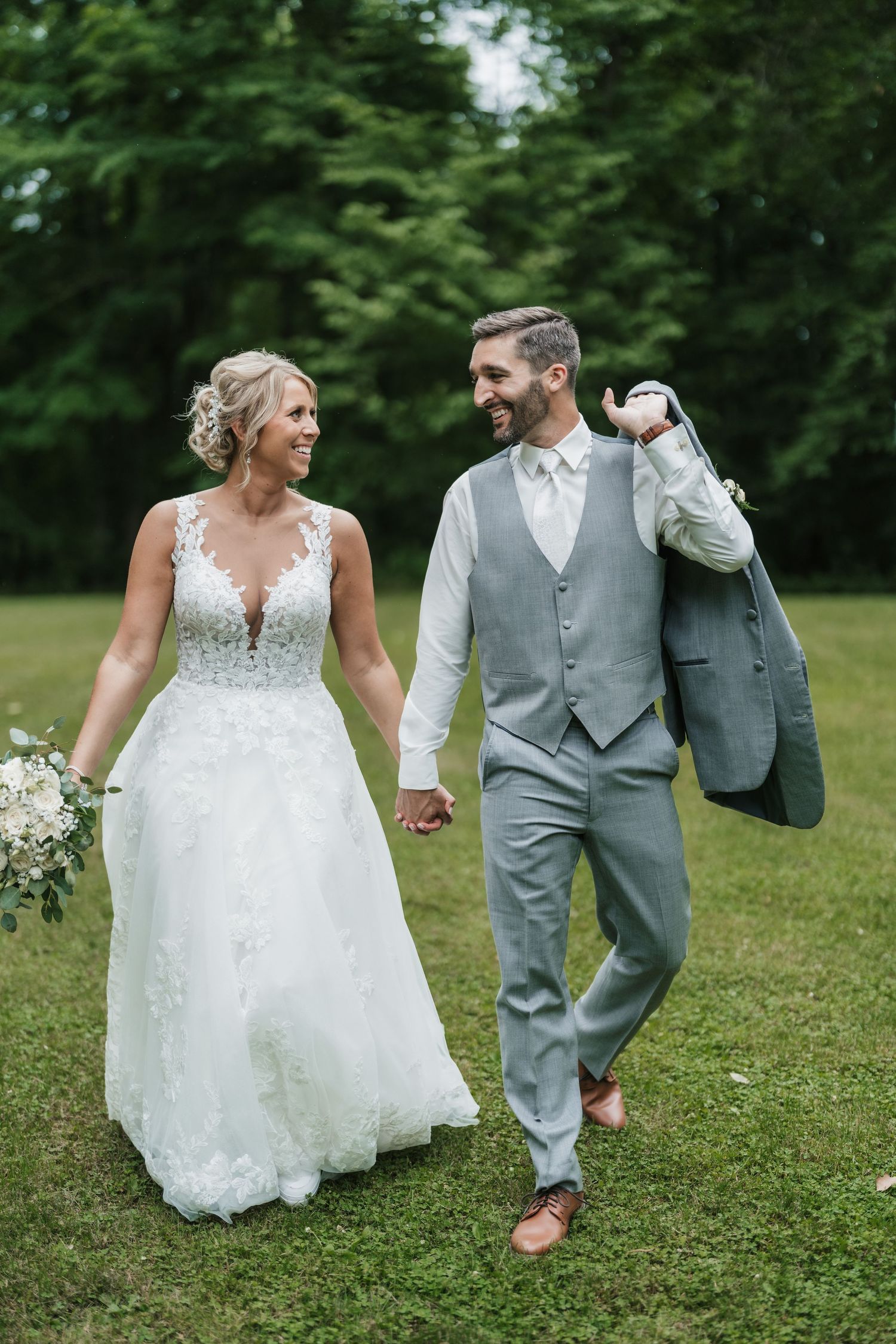 Newlyweds walk hand in hand through green grass with groom carrying suit jacket.