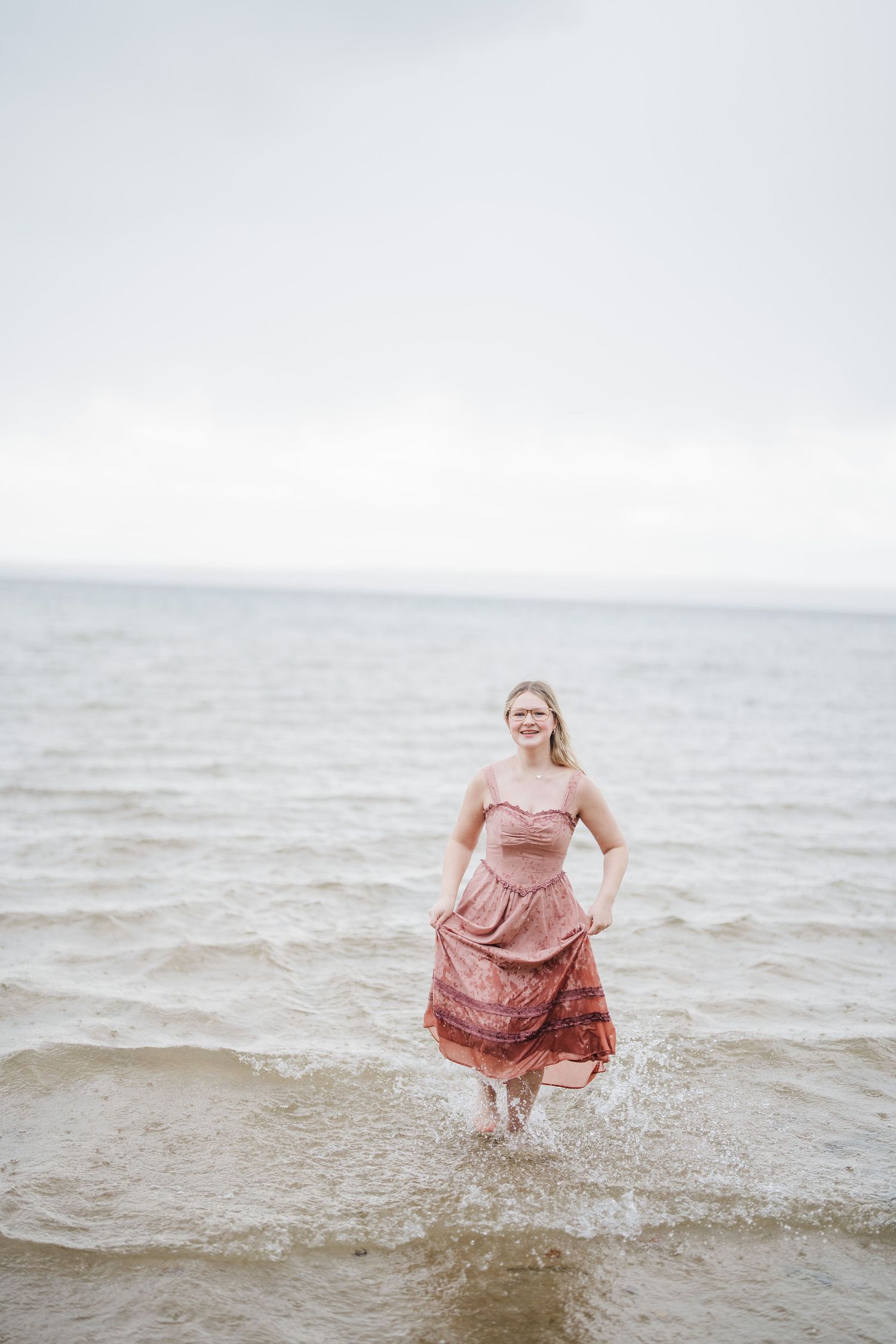 Someone in a flowing pink dress splashes joyfully in shallow ocean waters at sunset.