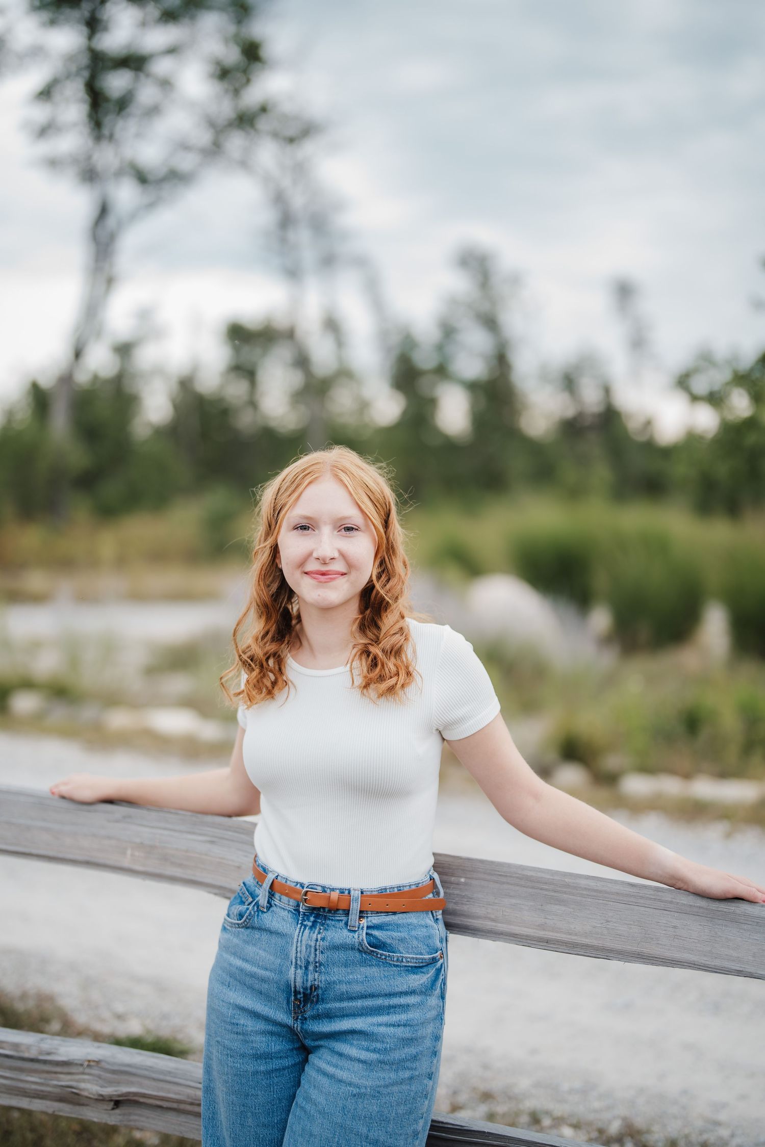 A person in a white t-shirt and jeans poses casually against a wooden railing with trees and nature in the background.