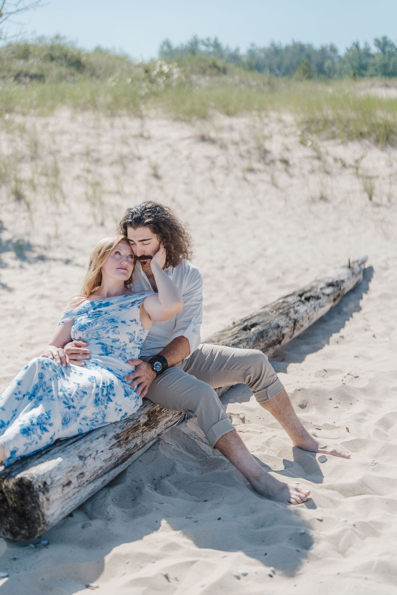 A couple shares an intimate moment while resting against a driftwood log on a sandy beach with dunes in the background.