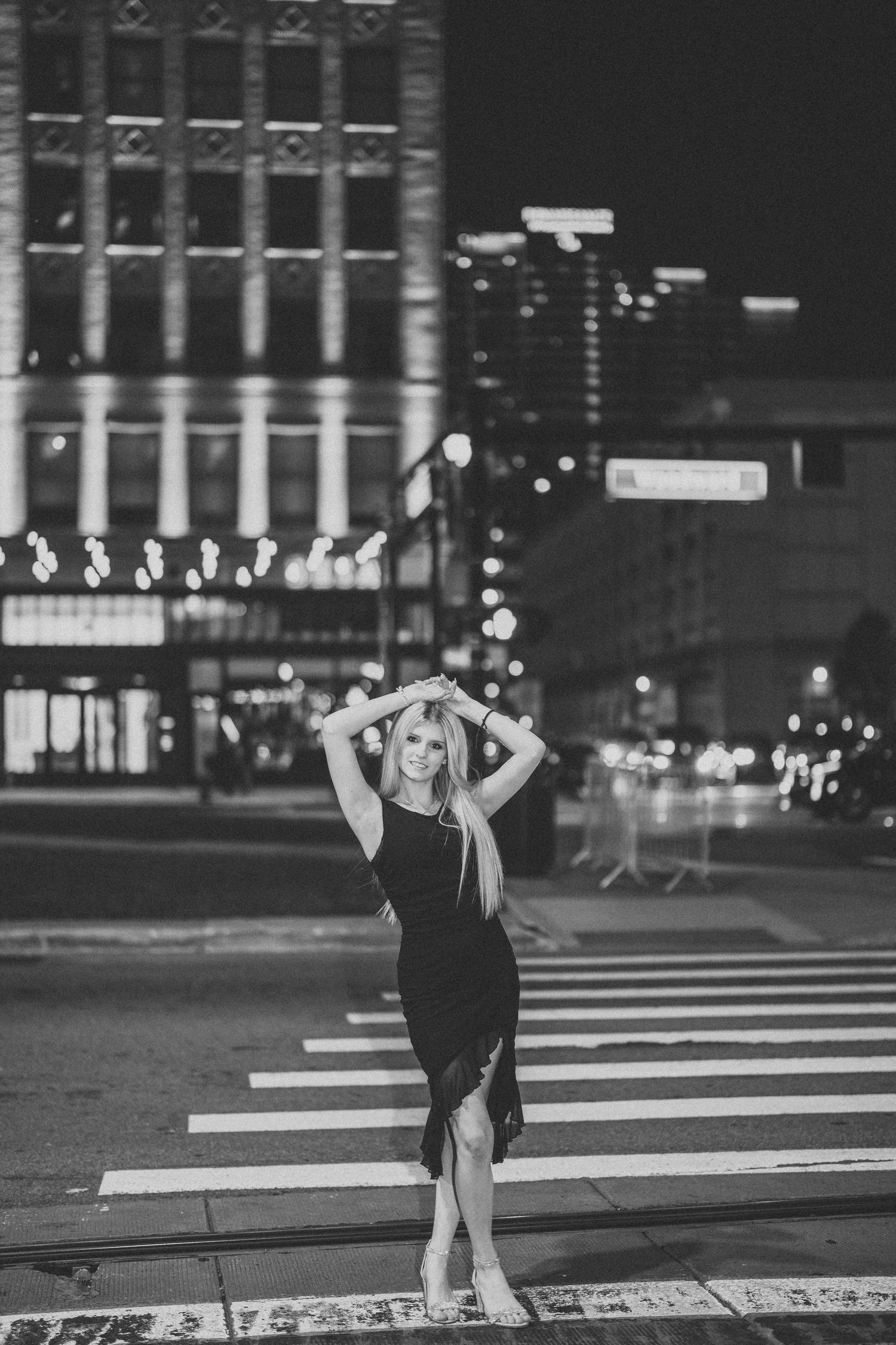 A person in a black dress poses on a crosswalk at night with city lights in the background.