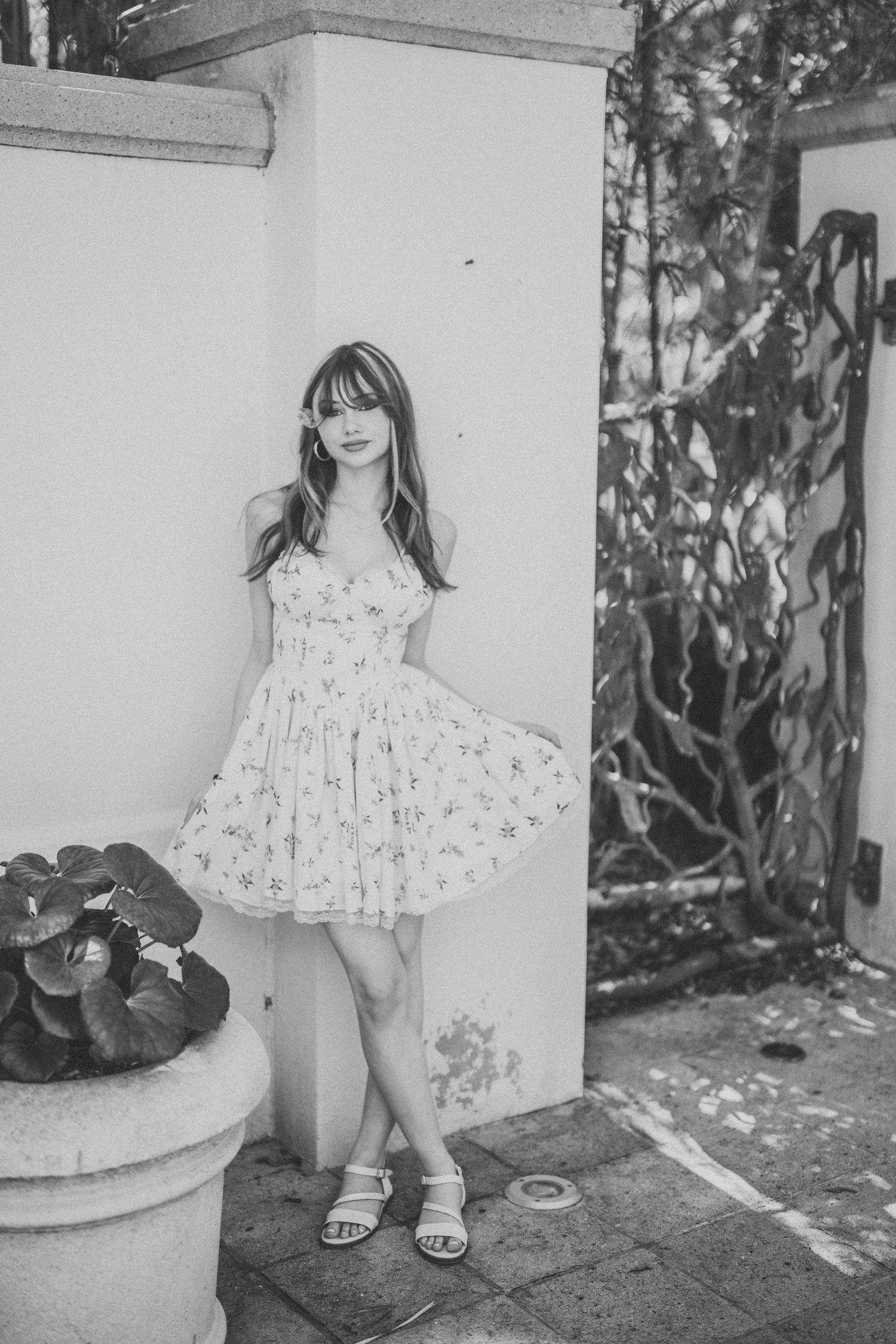 A black and white photo series showing a child in a floral dress posing against a white wall with potted plants nearby.
