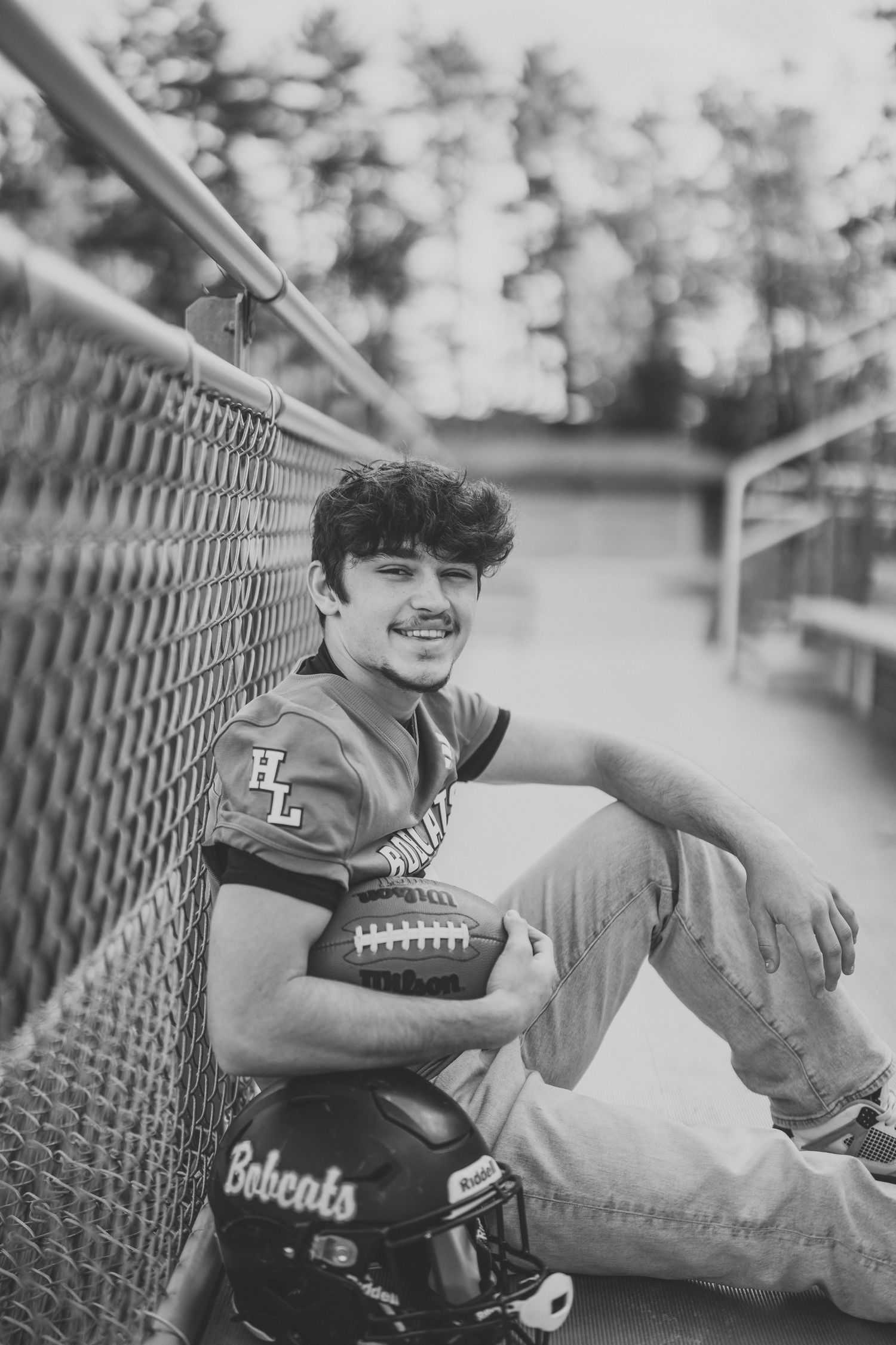 Black and white photo of athlete lounging on bleachers with football and helmet.