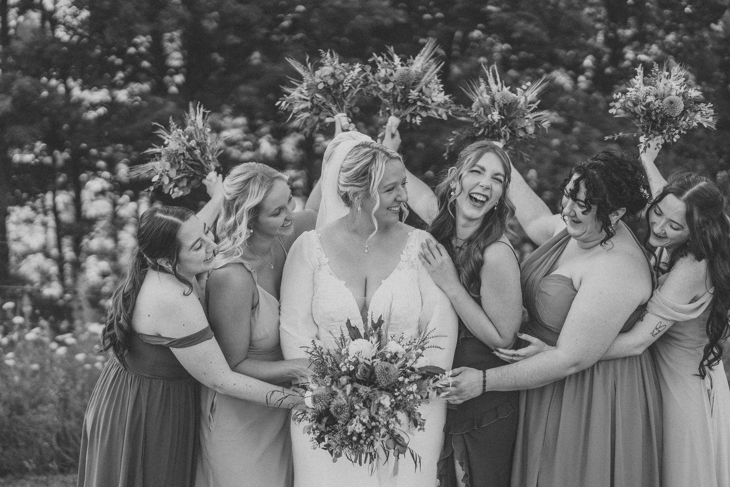 A joyful bridal party in gray dresses shares a candid moment outdoors in black and white photography.