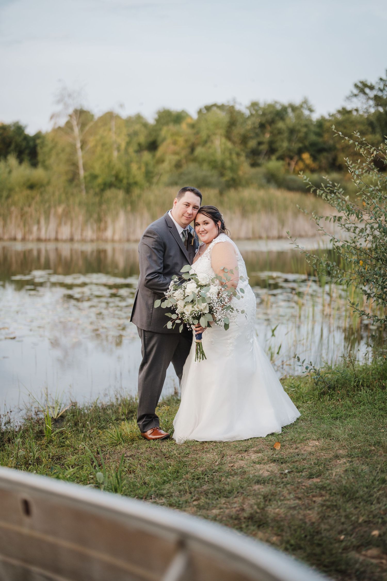 A couple poses together by a serene pond surrounded by autumn trees while holding a white floral bouquet.