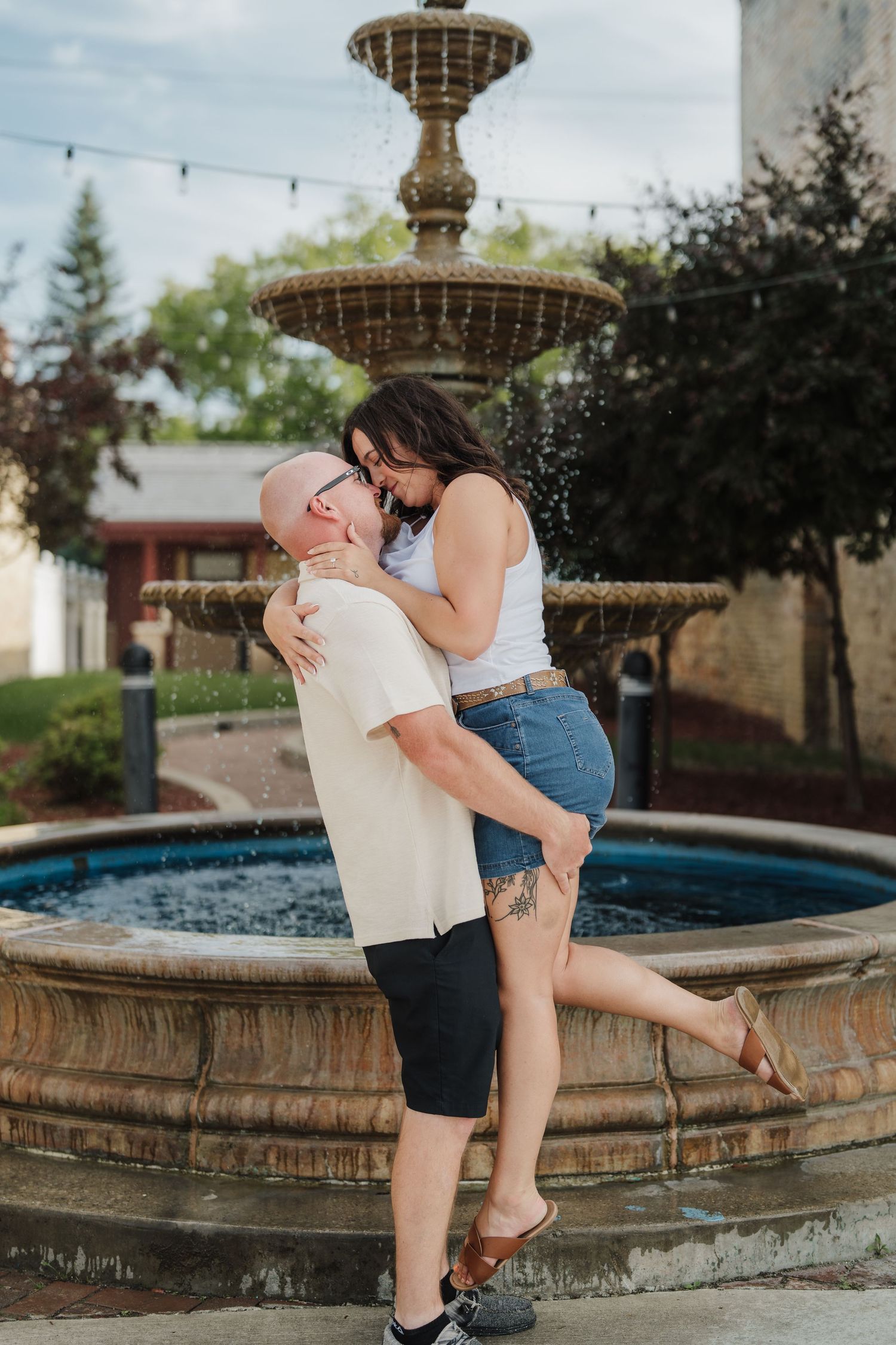 Couple embracing and kissing in front of a decorative stone fountain outdoors, woman lifted slightly off the ground.