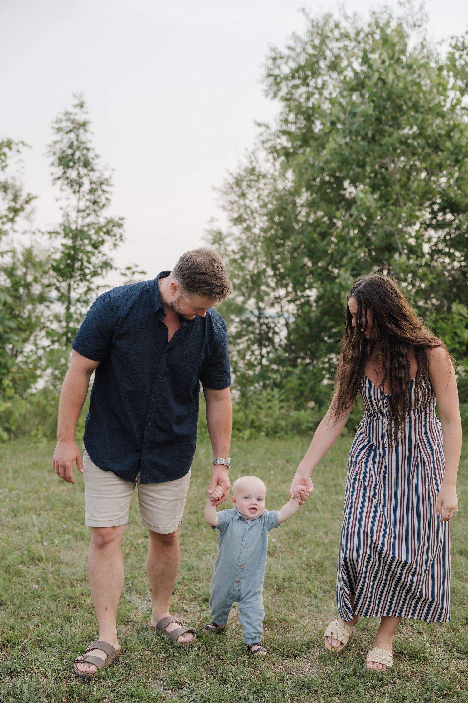 A young family walks outdoors, parents holding their baby's hands on a grassy field with green trees in the background.