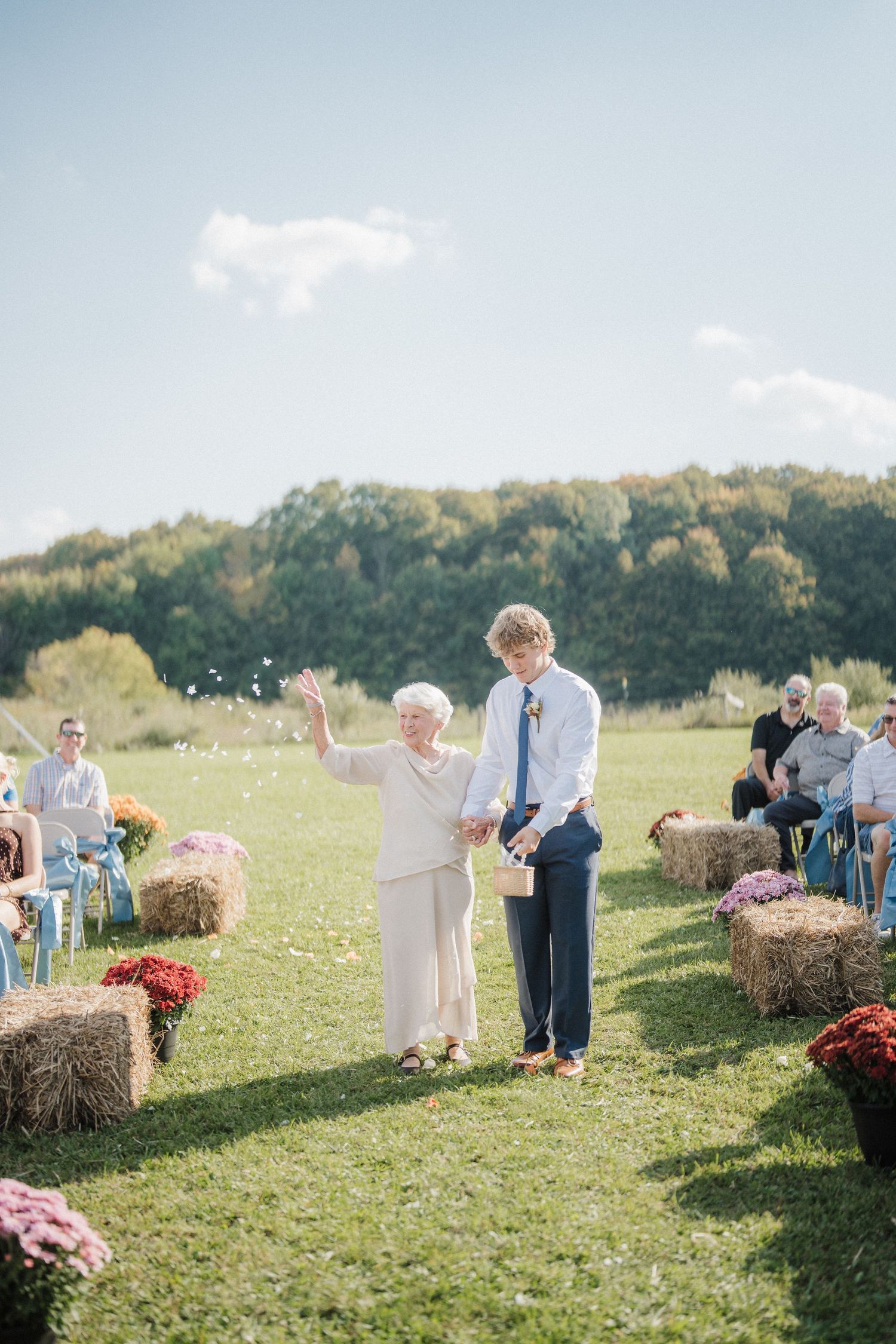 Wedding guests sit on hay bales in a sunny outdoor field decorated with flowers for a rustic countryside ceremony.