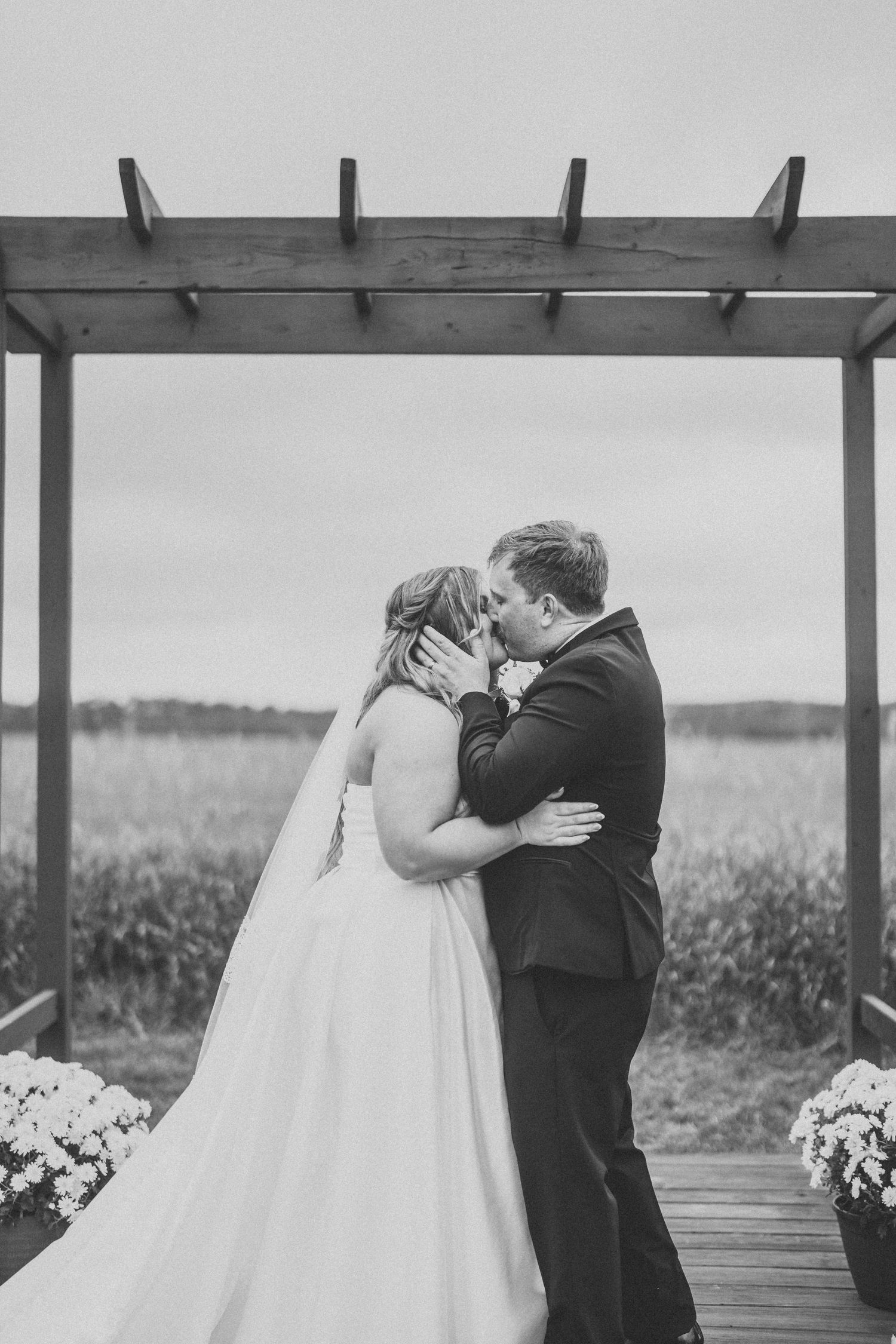 A romantic wedding moment captured in black and white under a wooden pergola with flower arrangements on the deck.