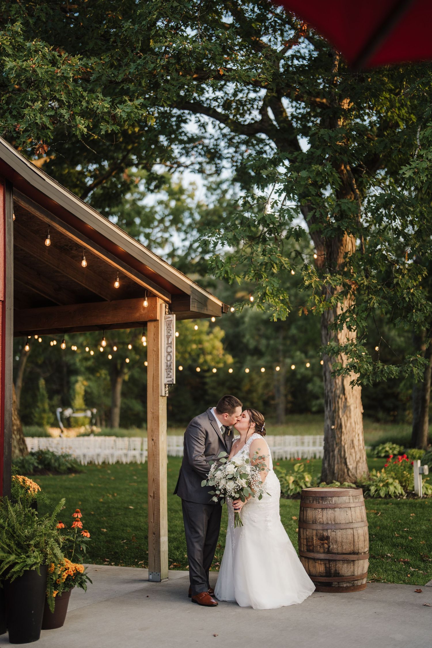 A romantic outdoor wedding scene with string lights and wooden barrels under a rustic wooden pergola at sunset.