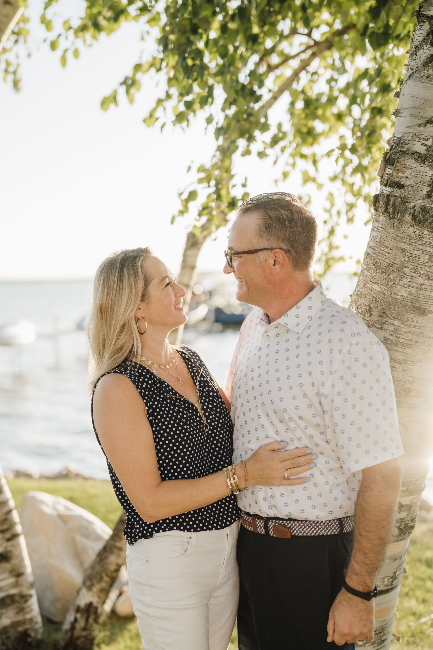 A couple shares an intimate moment under birch trees by the water's edge during golden hour.