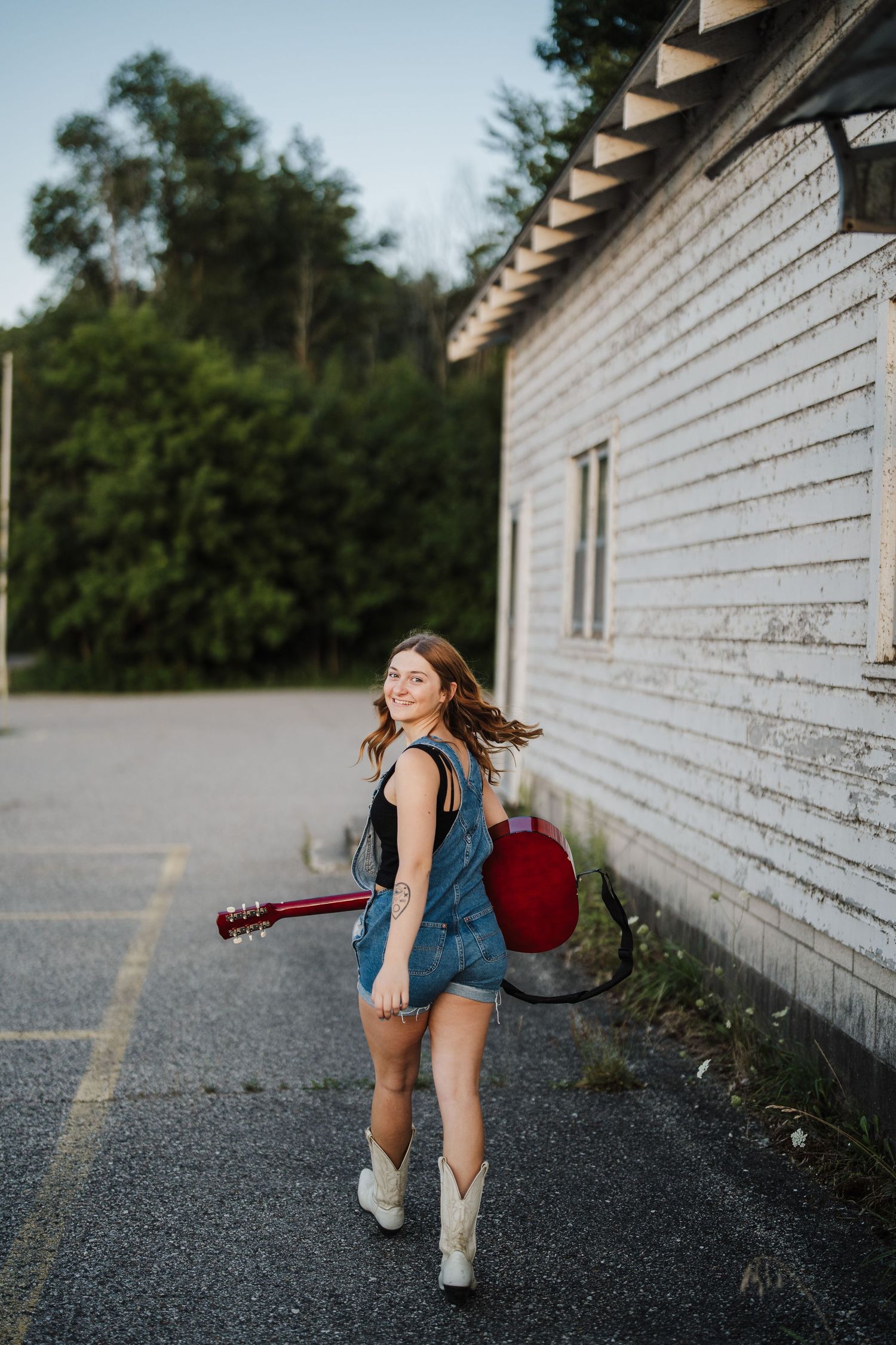 Figure in denim shorts and backpack poses playfully against white building wall at sunset.