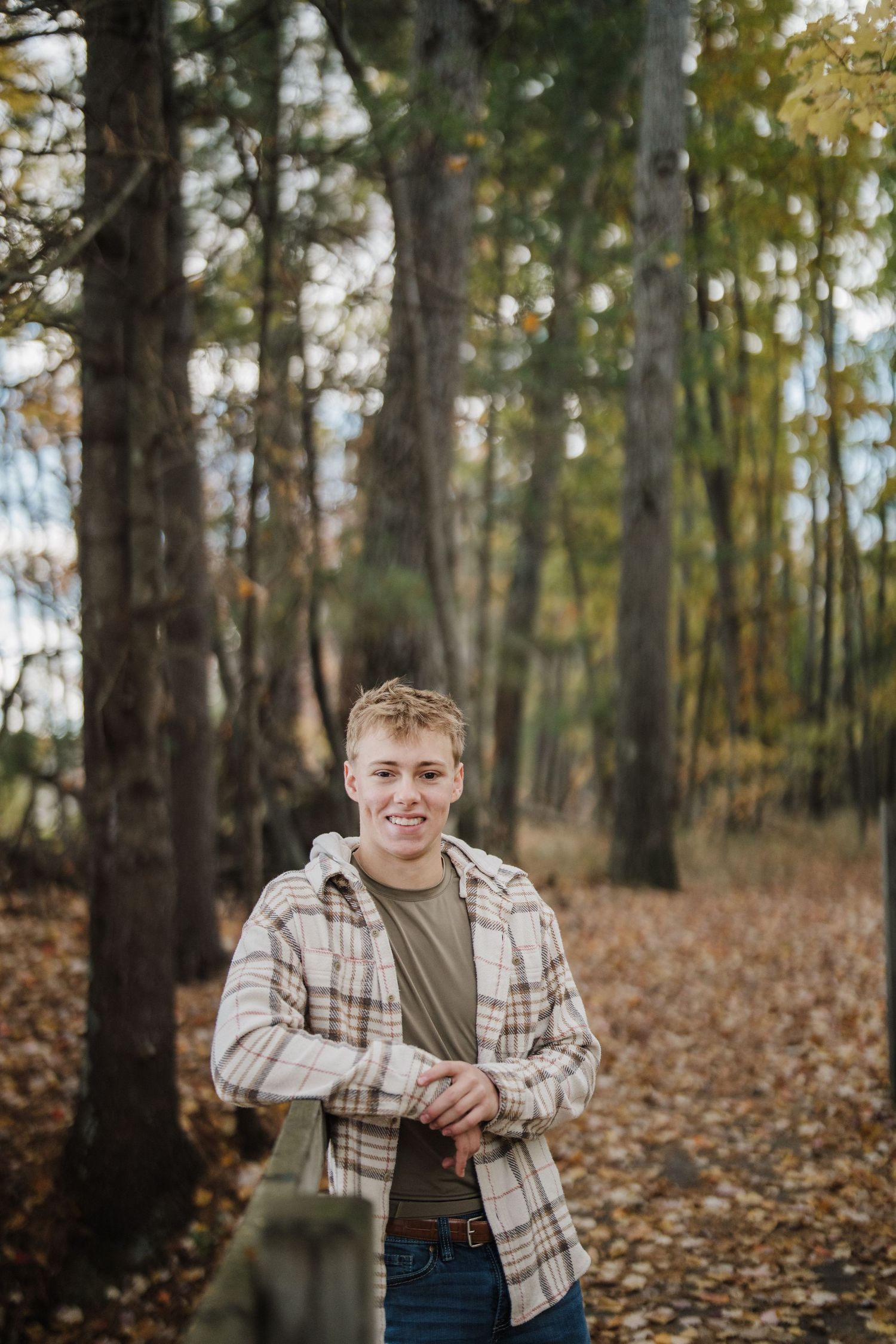 A person poses outdoors in autumn wearing a plaid shirt and olive shirt among fallen leaves and tall trees.